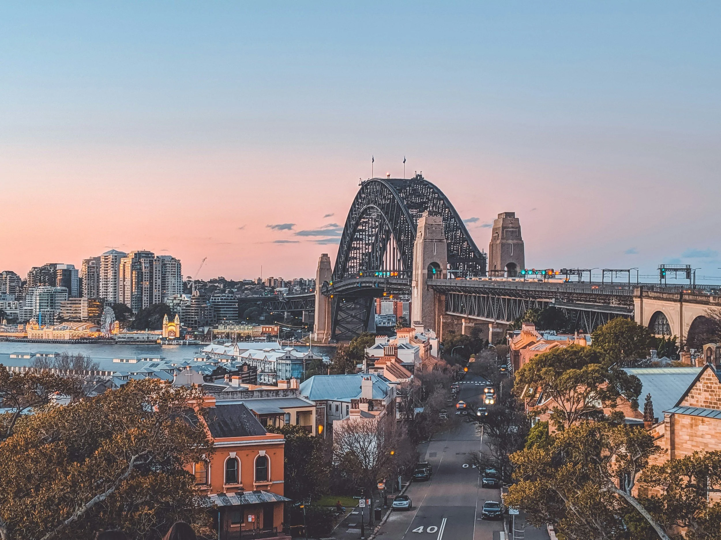Sydney Harbour Bridge at sunset, with city skyscrapers in the background and cars on the bridge.