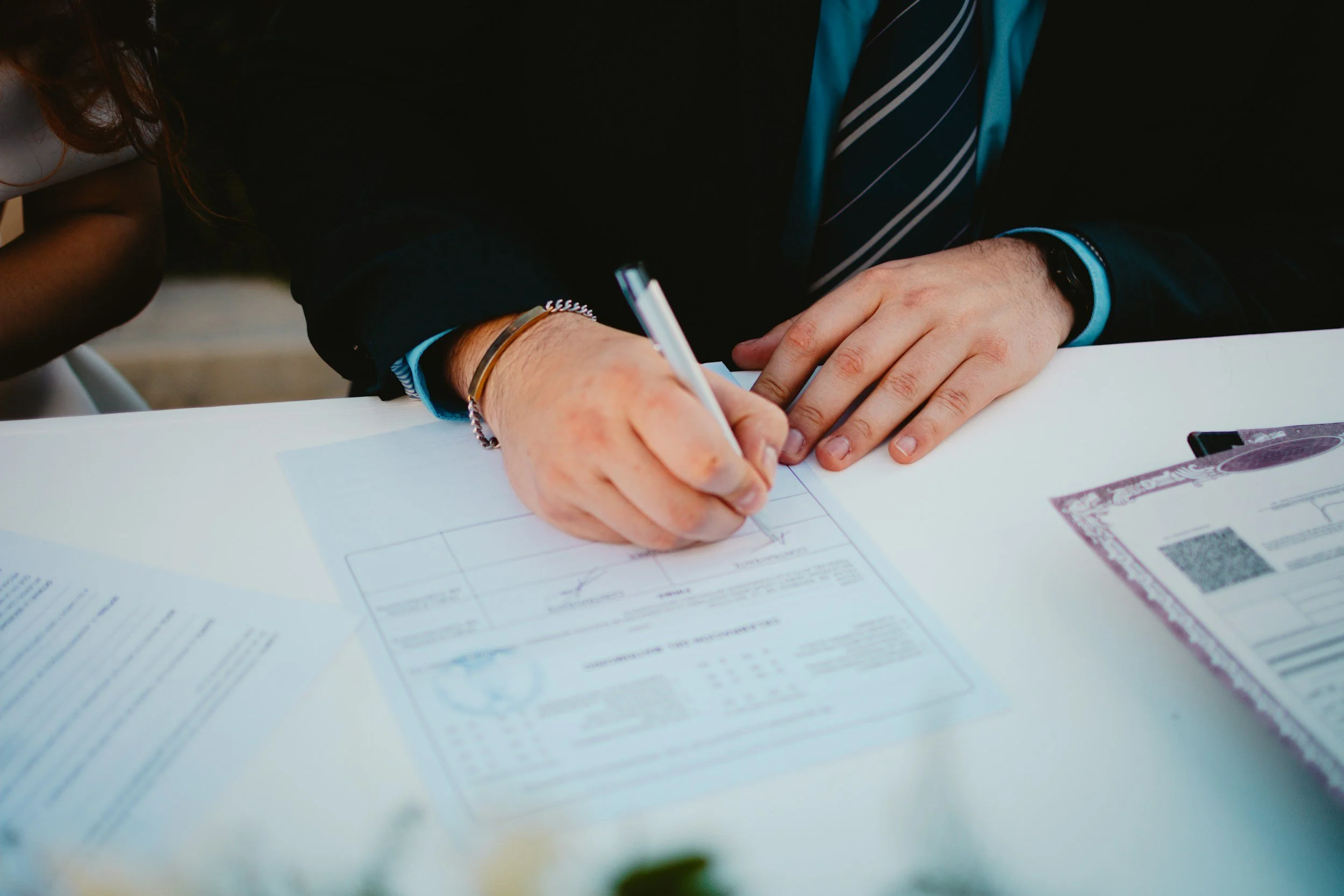 Close-up of a person in a black suit and striped tie signing papers on a white table, with US dollar bills in the foreground.