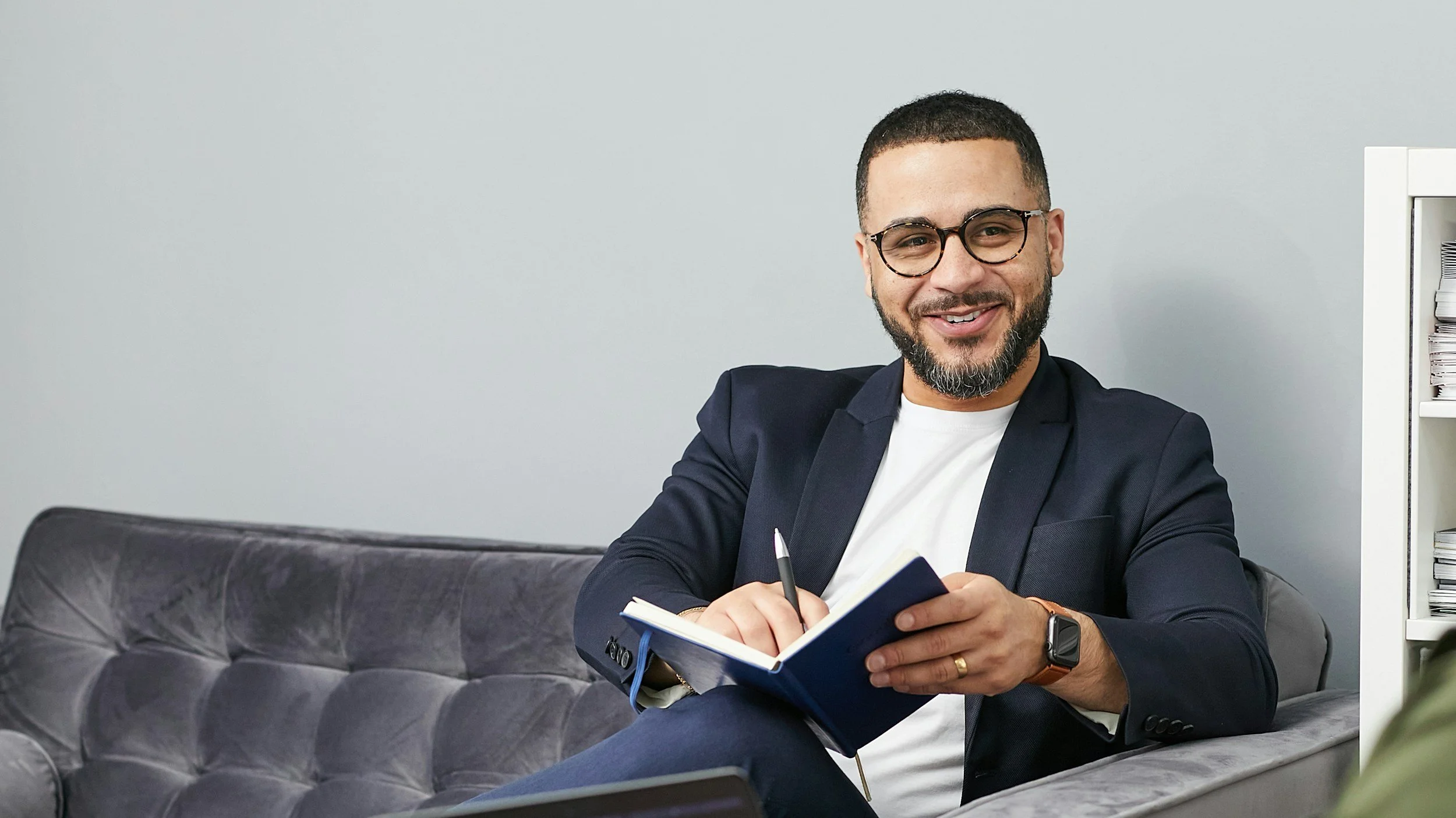 Smiling man with glasses and a beard sitting on a sofa with a notebook and pen, dressed in a dark blazer and white shirt, in an office setting.