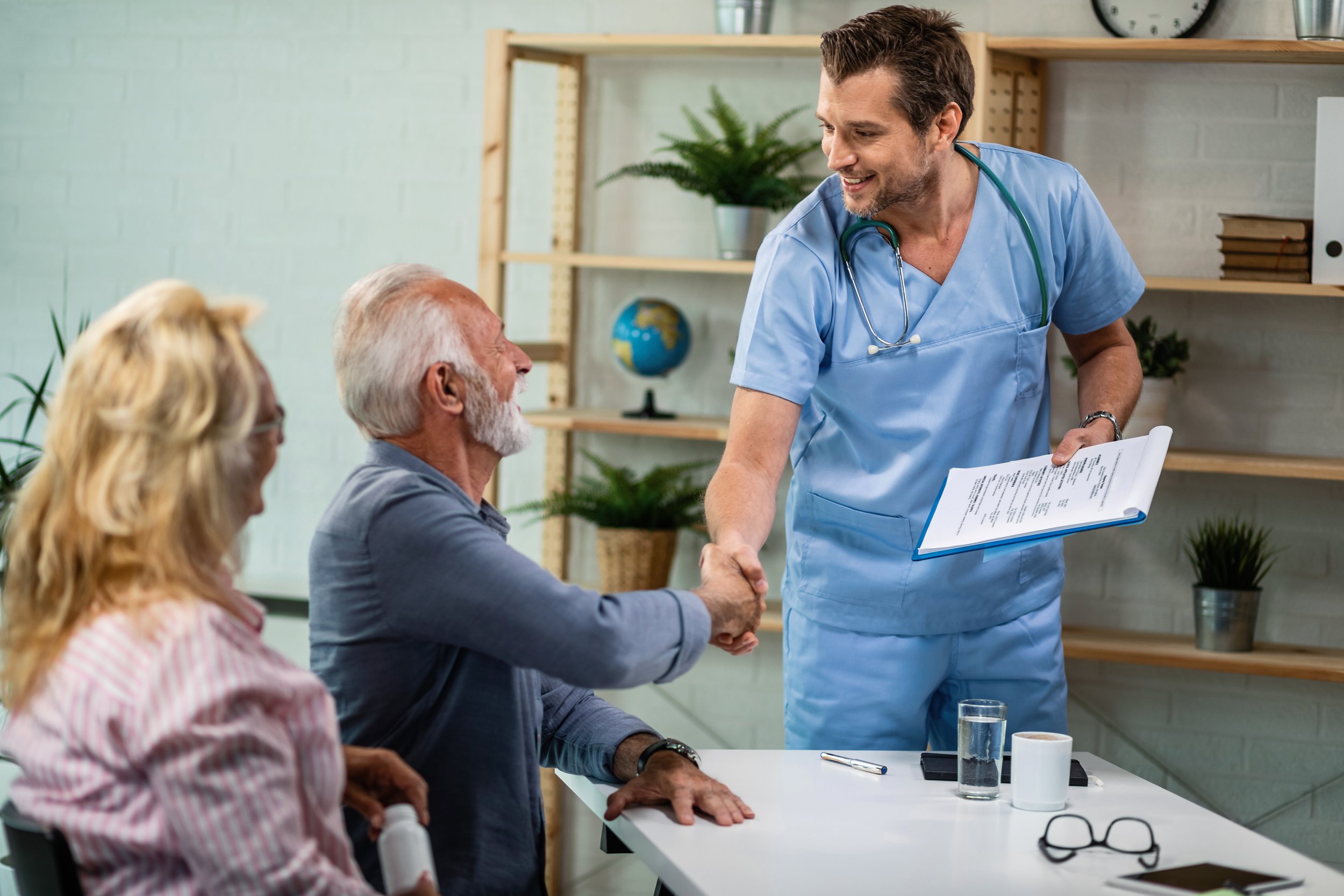 A healthcare professional in blue scrubs and a stethoscope shaking hands with an elderly man in a medical consultation room. Two other elderly individuals, a man and a woman, are sitting at a table, observing the handshake.