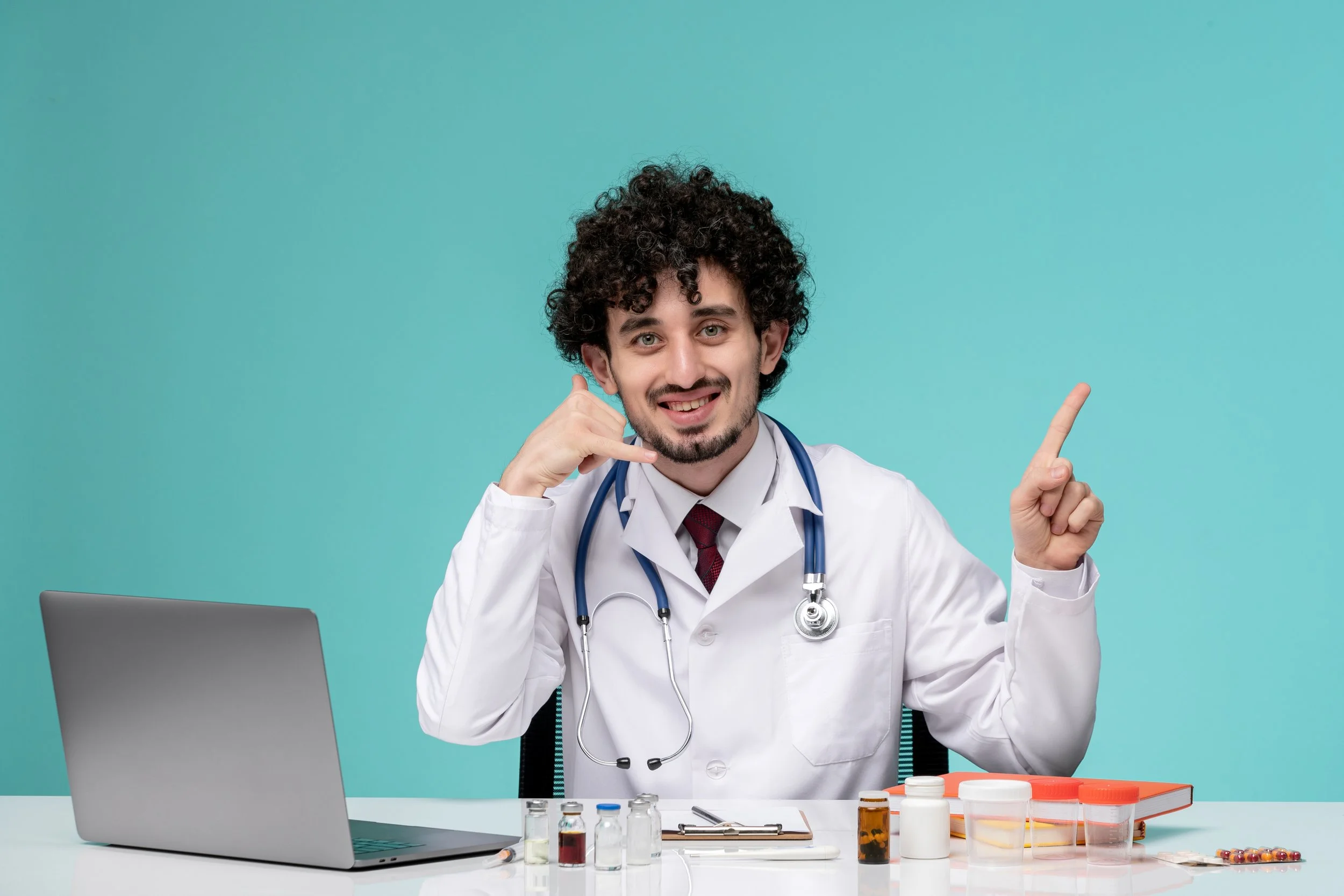 A young male doctor with curly dark hair, wearing a white lab coat and stethoscope, is sitting at a desk covered with medication bottles and medical supplies, smiling and pointing upward.