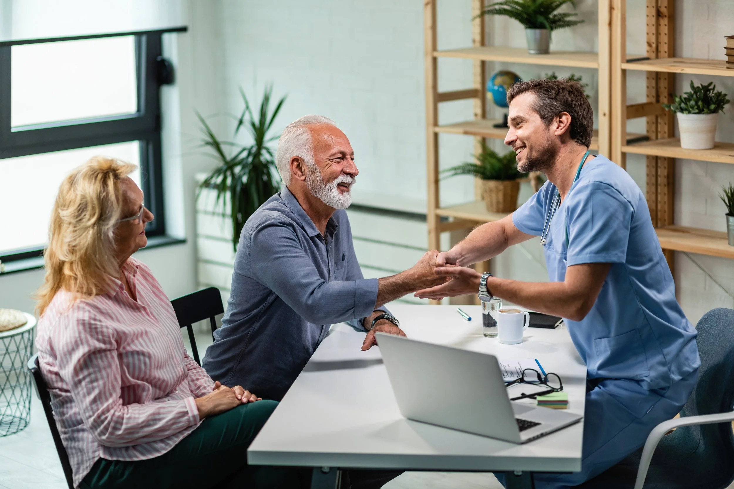 A healthcare professional in blue scrubs shaking hands with an elderly man, with an elderly woman and a glass of water and coffee mug on a white table, in a bright office with bookshelves and plants.