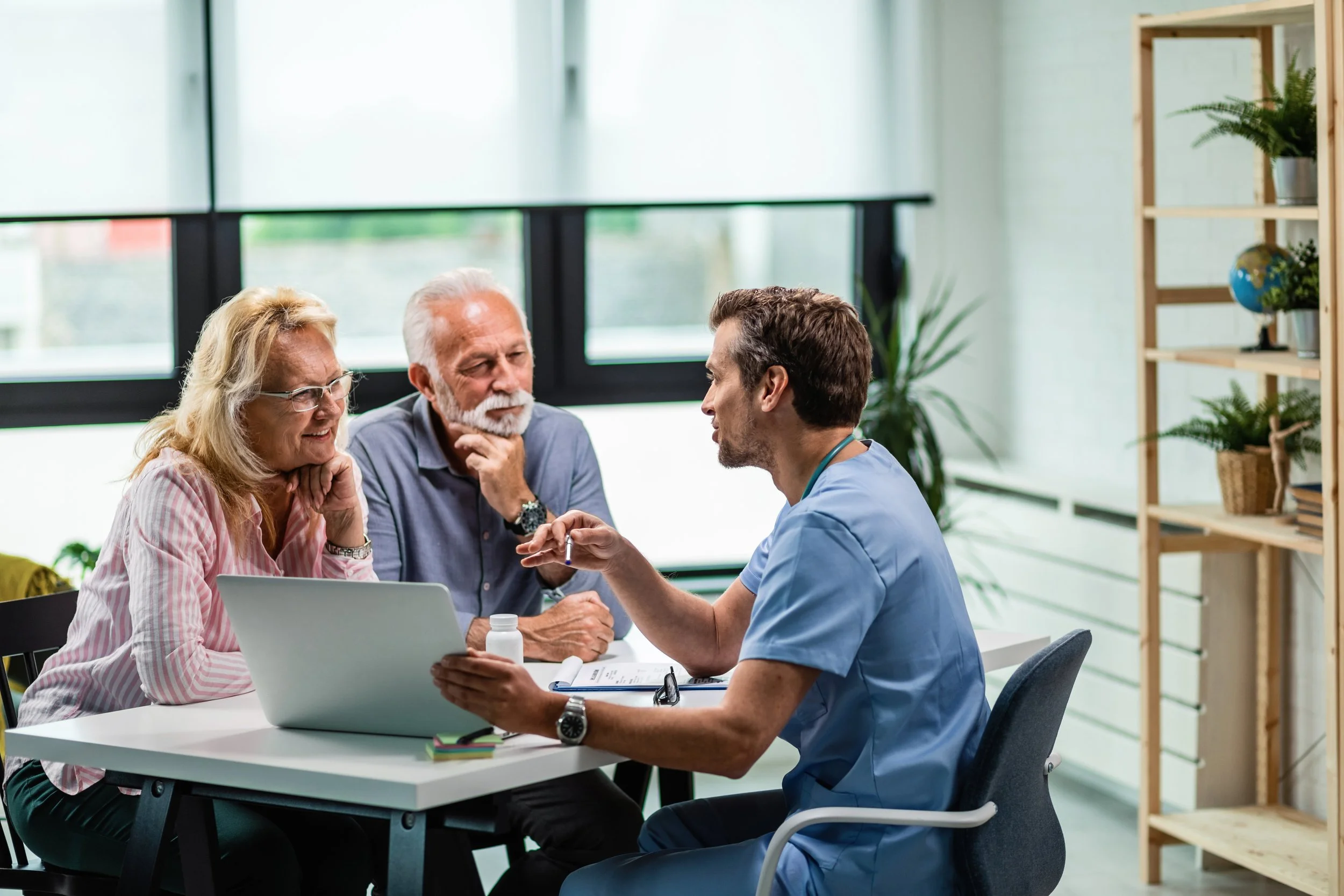 A doctor consulting with an elderly couple in a bright, modern office with large windows, plants, and a wooden bookshelf.