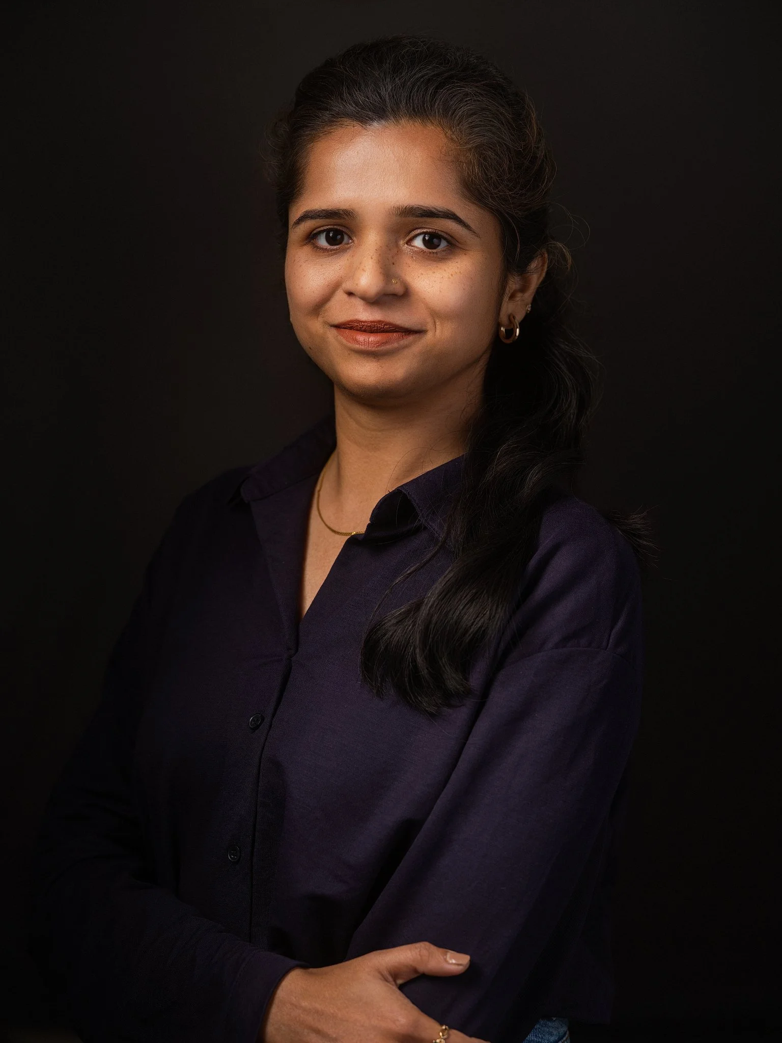 Portret van een jonge vrouw met lange, donkere haar in een donkere blouse, kijkend naar de camera, op een zwarte achtergrond.
