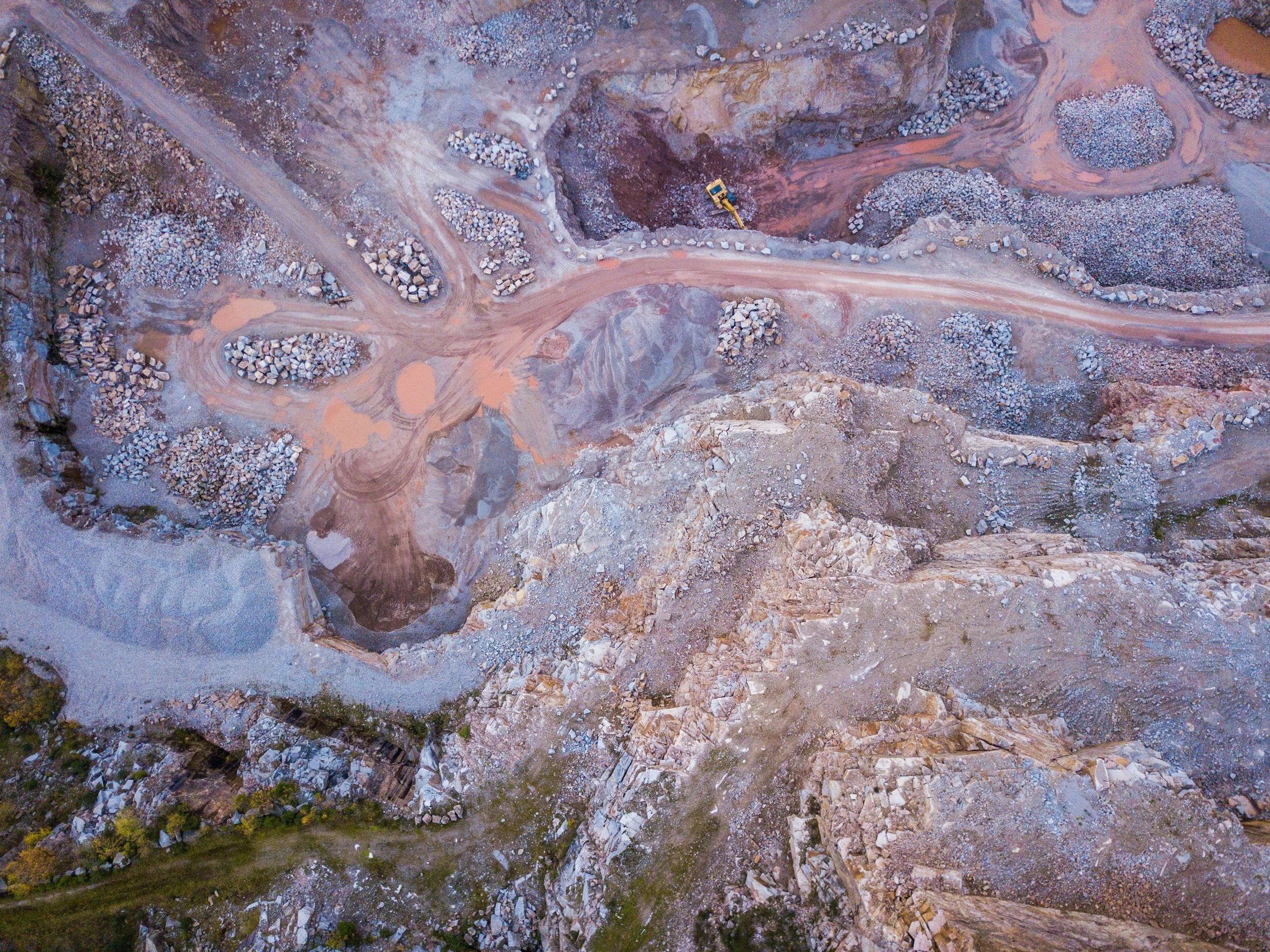 Aerial view of a quarry with winding dirt roads, mounds of rocks, and a yellow excavator.