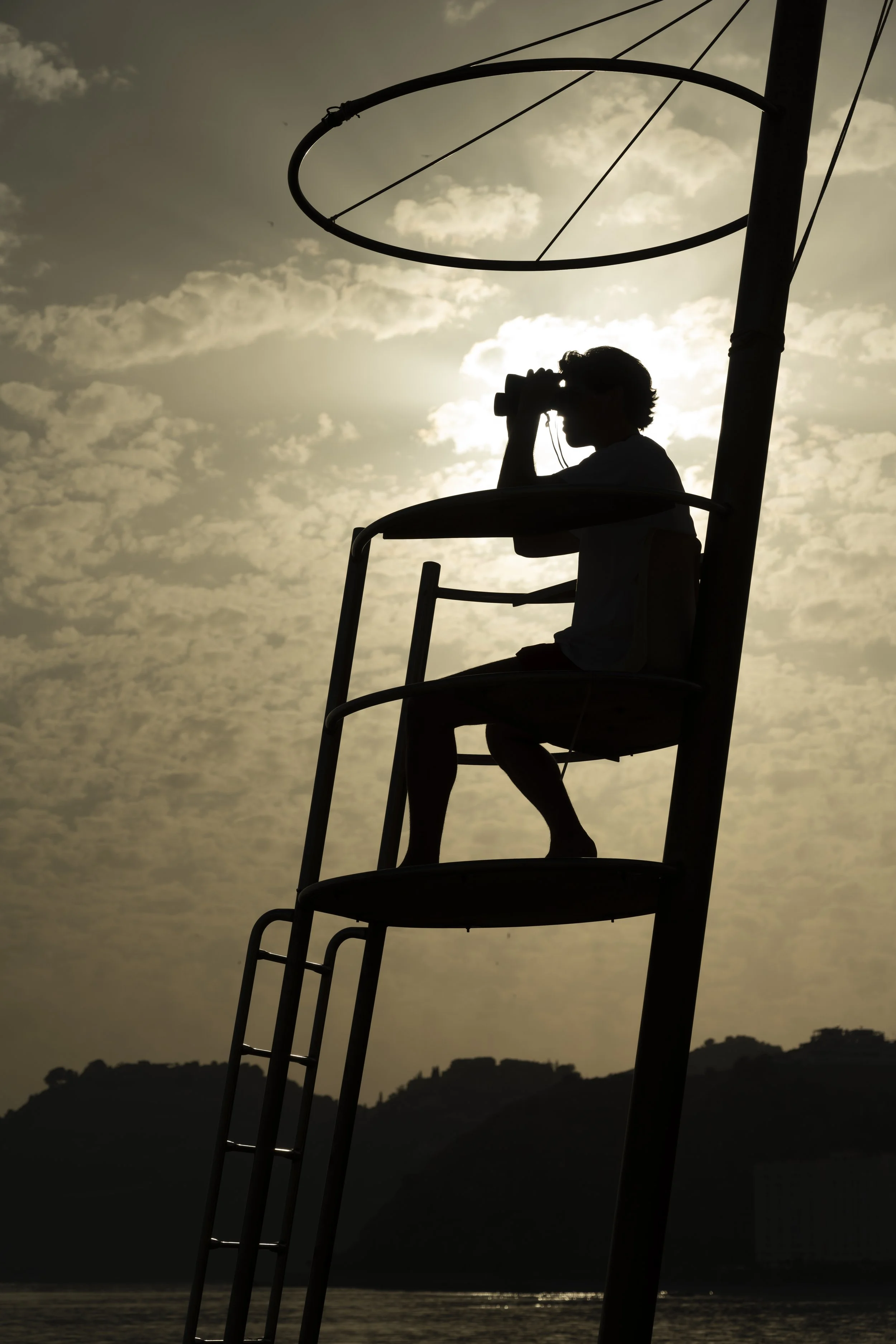 Silhouette of a boy sitting on a tall lifeguard chair, looking through binoculars, with the sun setting behind him and clouds in the sky.