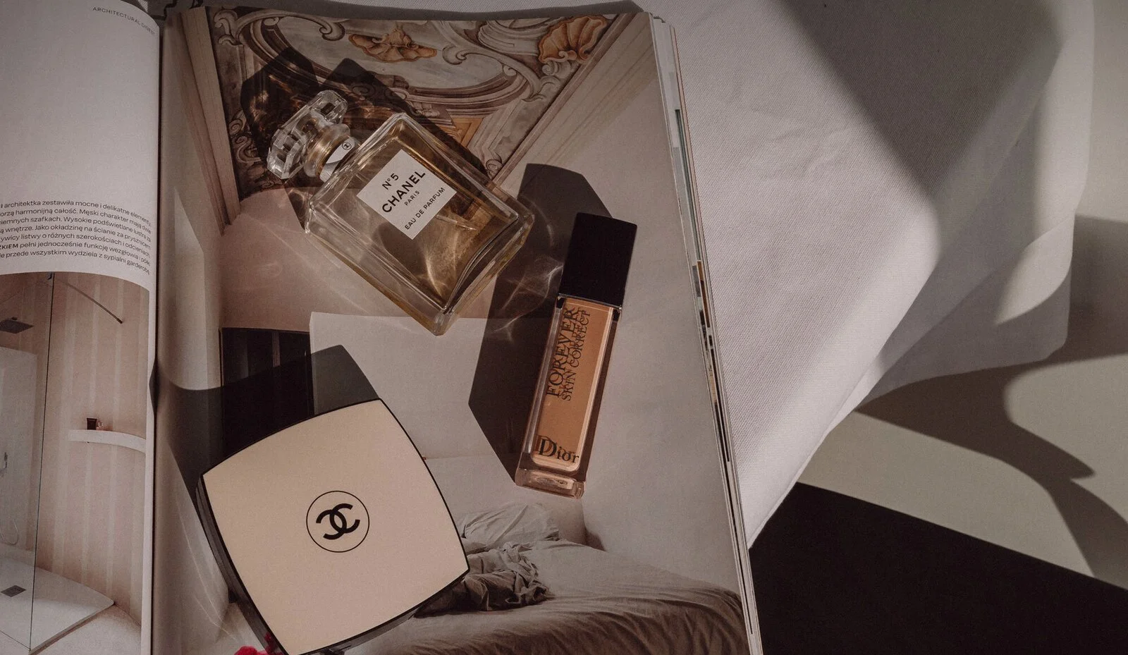 A magazine page displaying various designer brands and perfumes, including Chanel N°5 Eau de Parfum, Dior, and Chanel makeup compact, with a bed and a bedside table partially visible in the background.