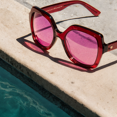 Pink sunglasses with purple lenses resting on a poolside concrete surface.