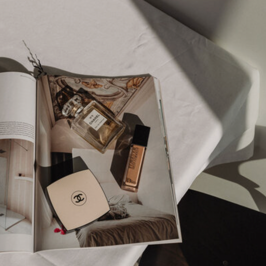 Open magazine on a white surface displaying a Chanel box, a bottle of Chanel perfume, and a bottle of foundation, with a soft light casting shadows.