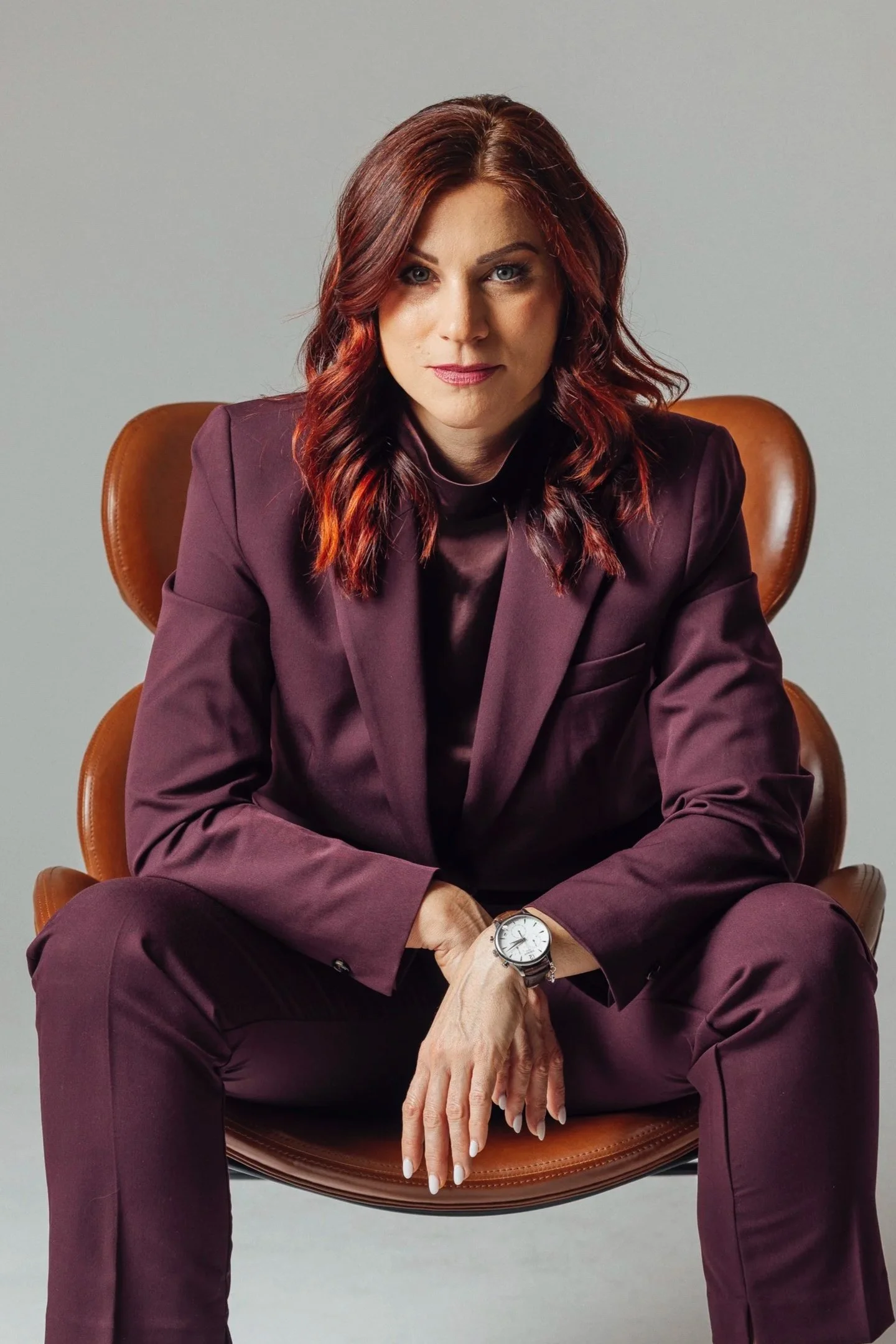 A woman with red hair wearing a dark purple suit, sitting on a brown leather chair against a plain gray background.