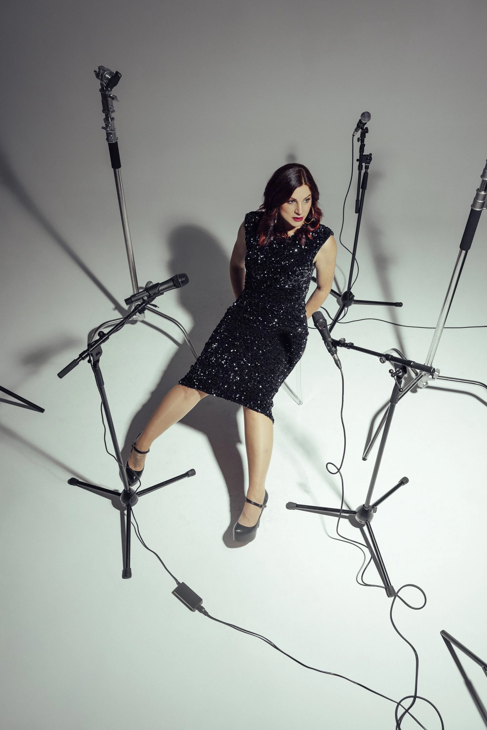 A woman in a black, sparkling dress sitting on a transparent chair surrounded by multiple microphones on stands in a studio setting.