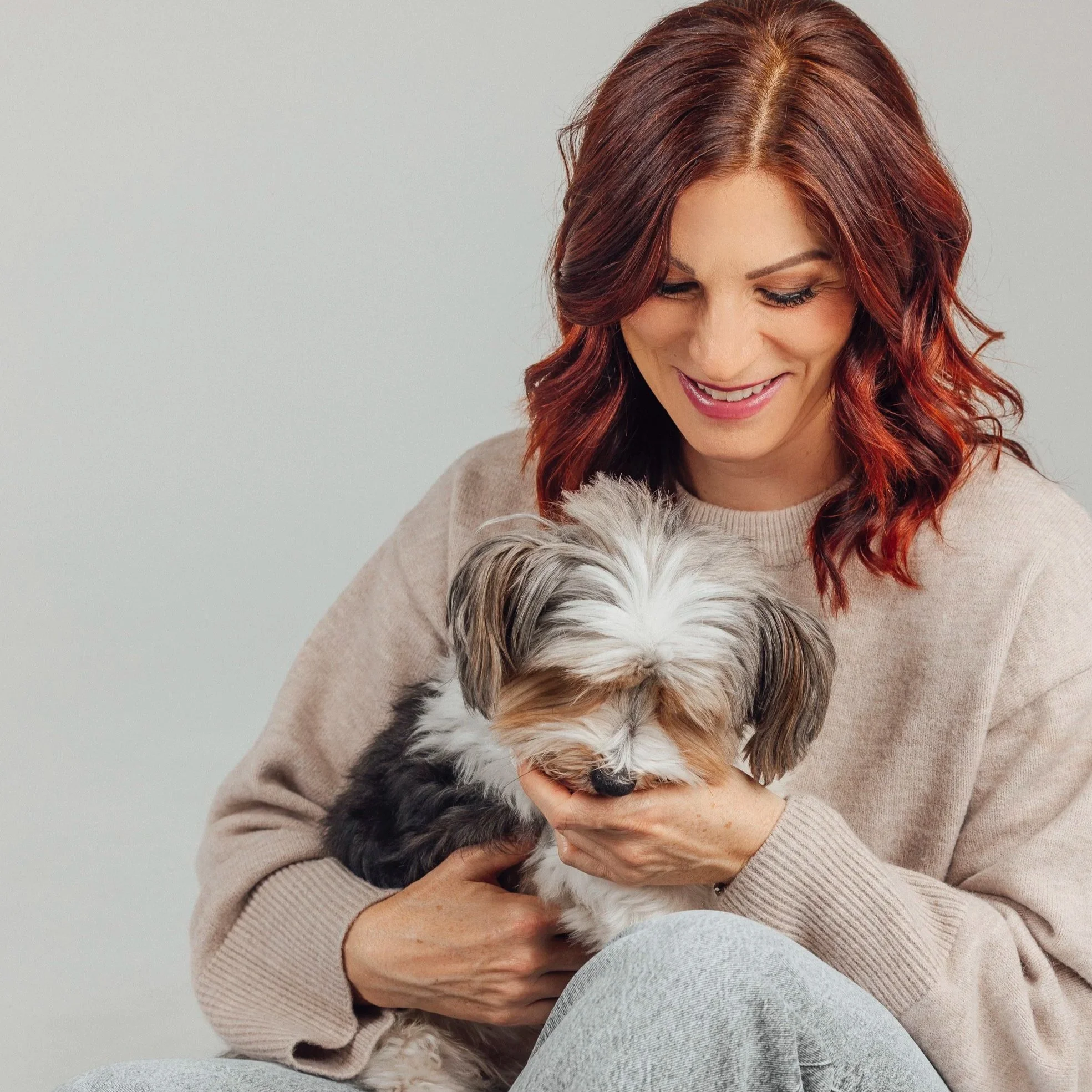 A woman with red hair smiling while holding a small fluffy dog with gray, white, and brown fur.
