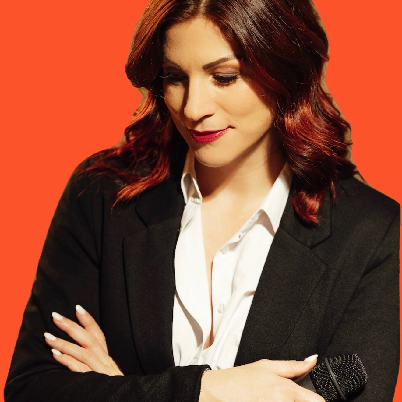 A woman with red hair and fair skin, wearing a black blazer and white shirt, looking down and crossing her arms, with a microphone in front of her, set against an orange background.