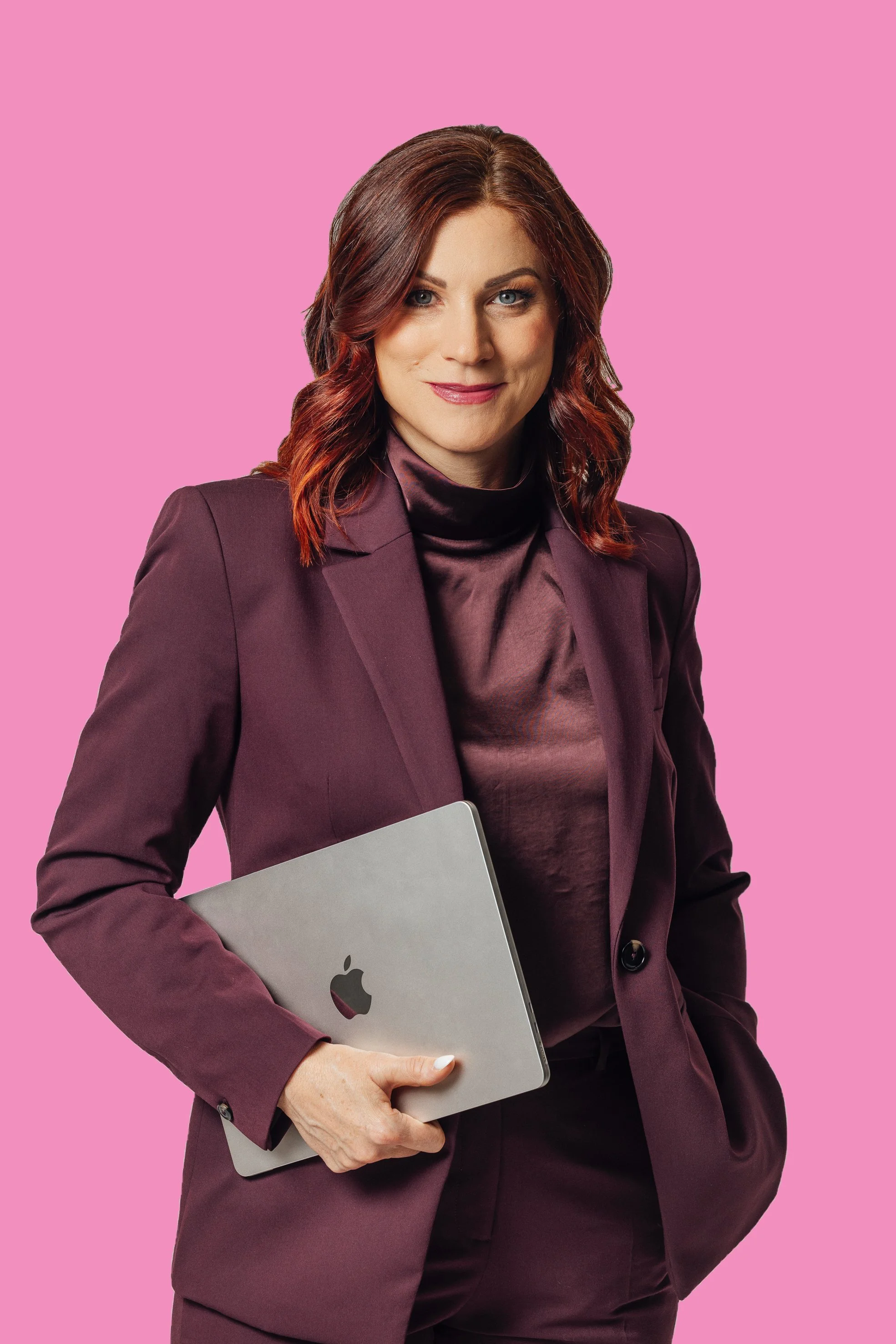 A woman with burgundy hair, wearing a maroon suit and holding a silver MacBook, standing against a pink background.