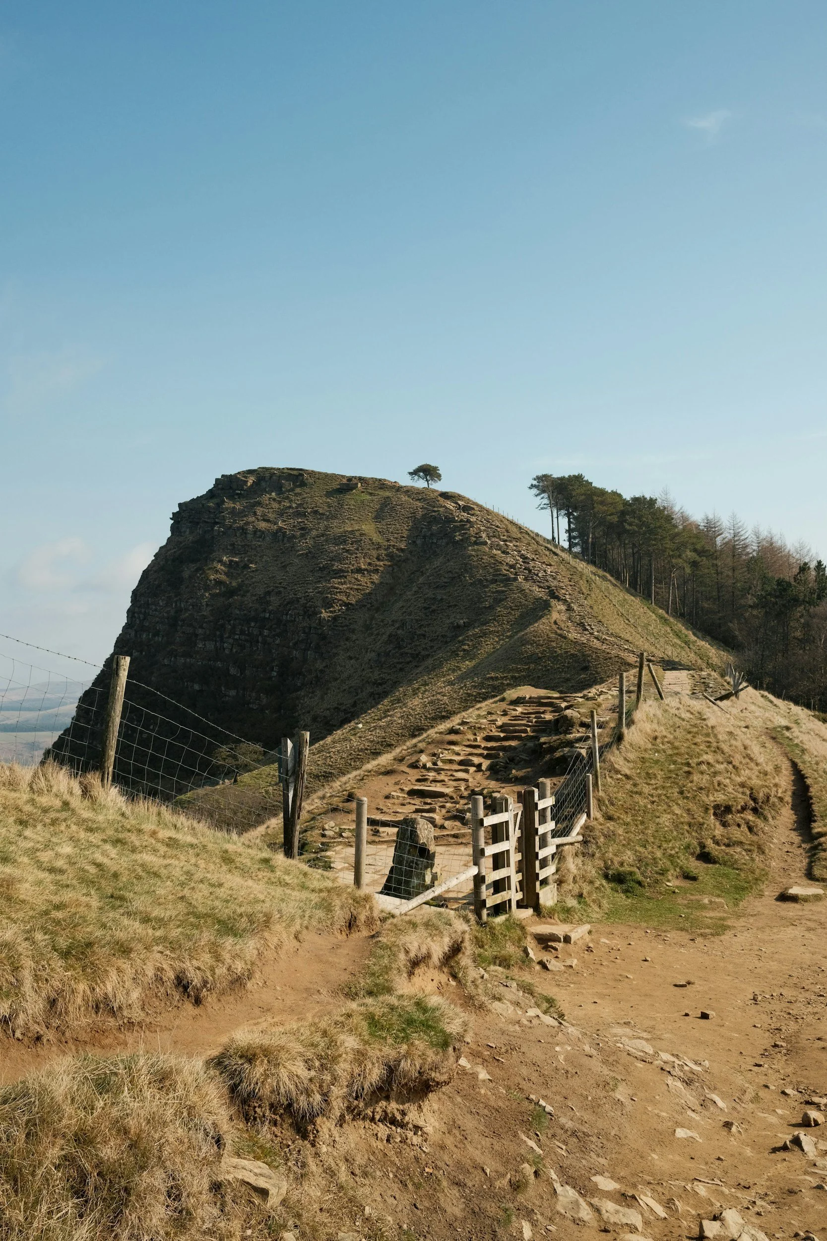 Hilly landscape with a dirt path, wooden fencing, and a prominent rock formation with a few trees on top under a blue sky.