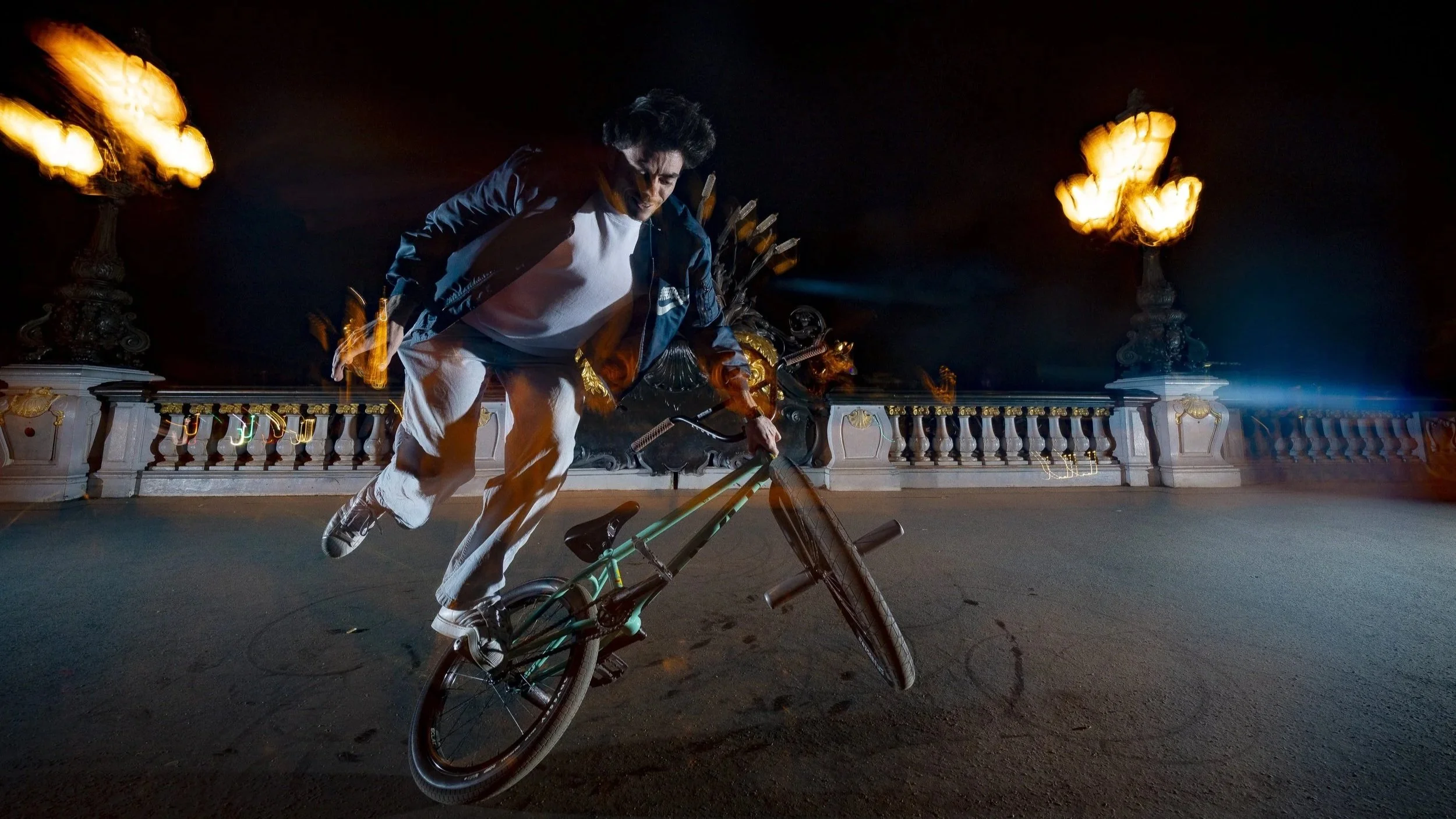 Homme faisant du BMX sur le pont neuf de Paris la nuit, avec des lumières floues dans le ciel.