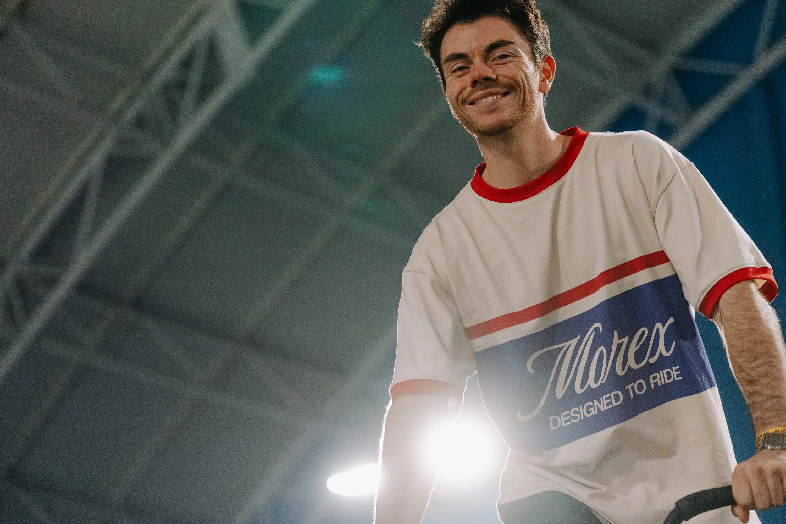 Un homme souriant portant un t-shirt blanc avec des bandes rouges et bleues, debout dans un environnement intérieur avec une structure métallique en arrière-plan, éclairé par une lumière forte derrière lui.