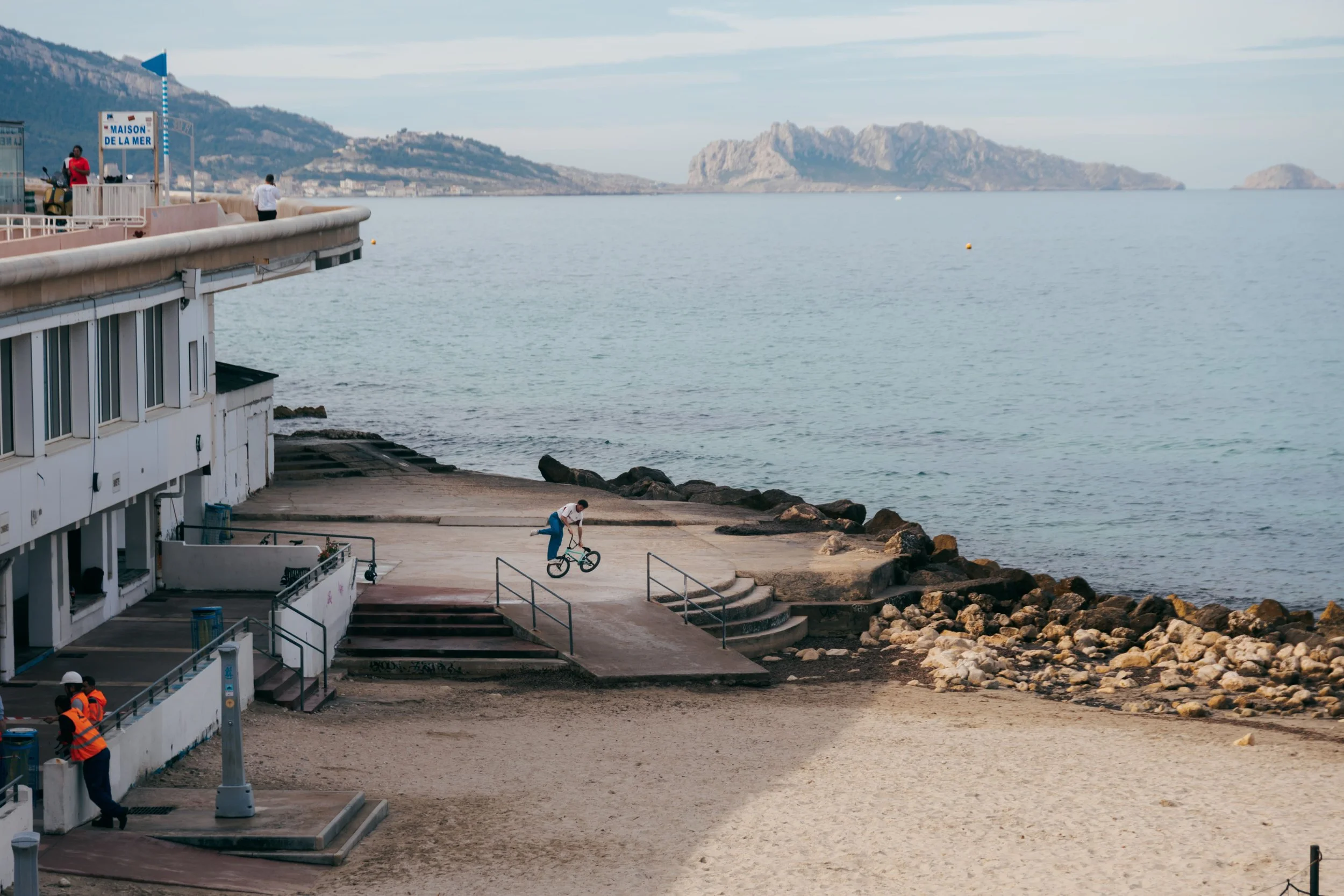 Vue large du spot de tournage au bord de la plage à Marseille, rider de BMX flat en trick face à la mer