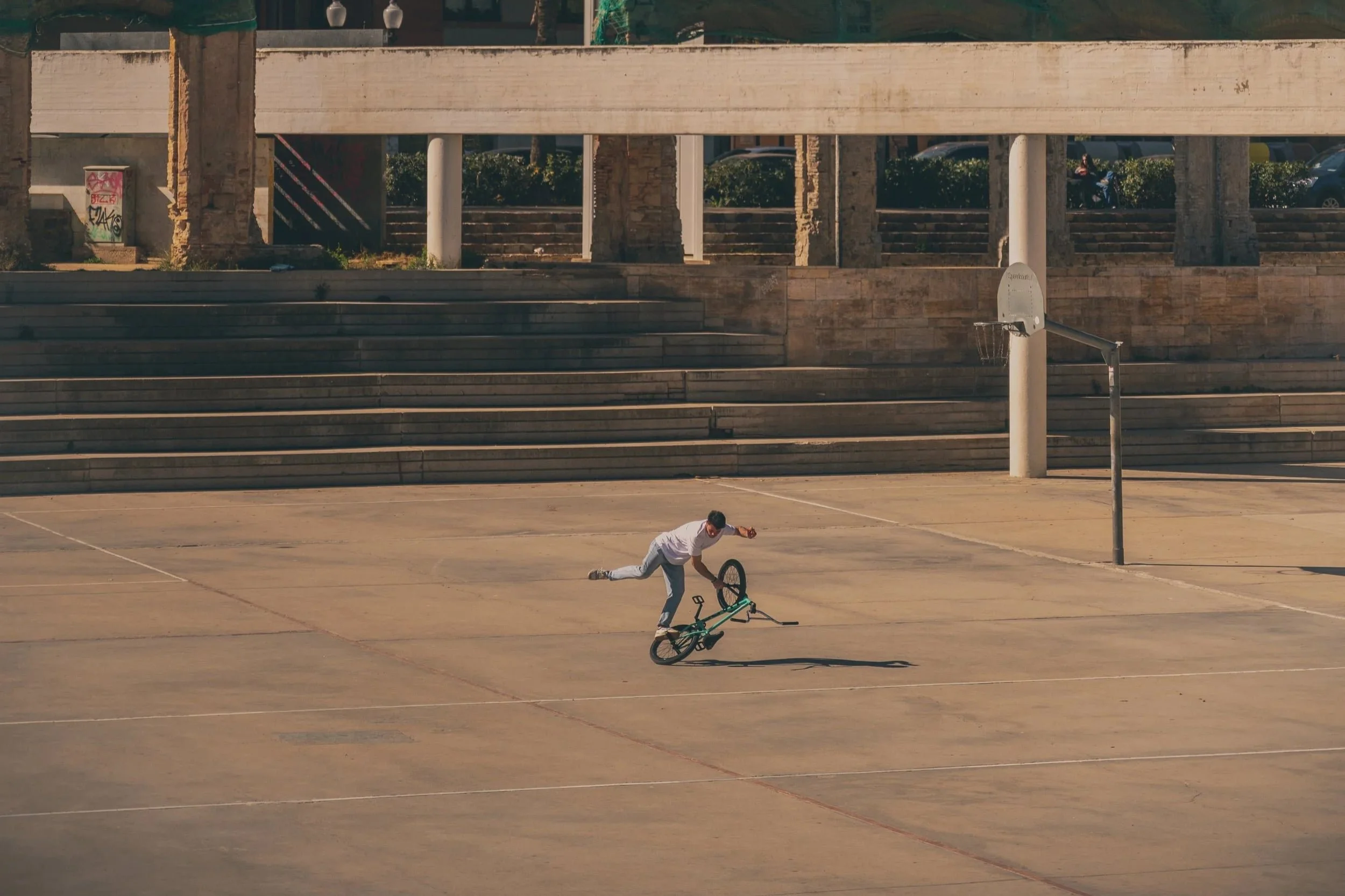Un garçon en train de faire un backflip sur son vélo dans un terrain de basketball vide, avec des gradins en pierre et une structure de basket-ball en arrière-plan.
