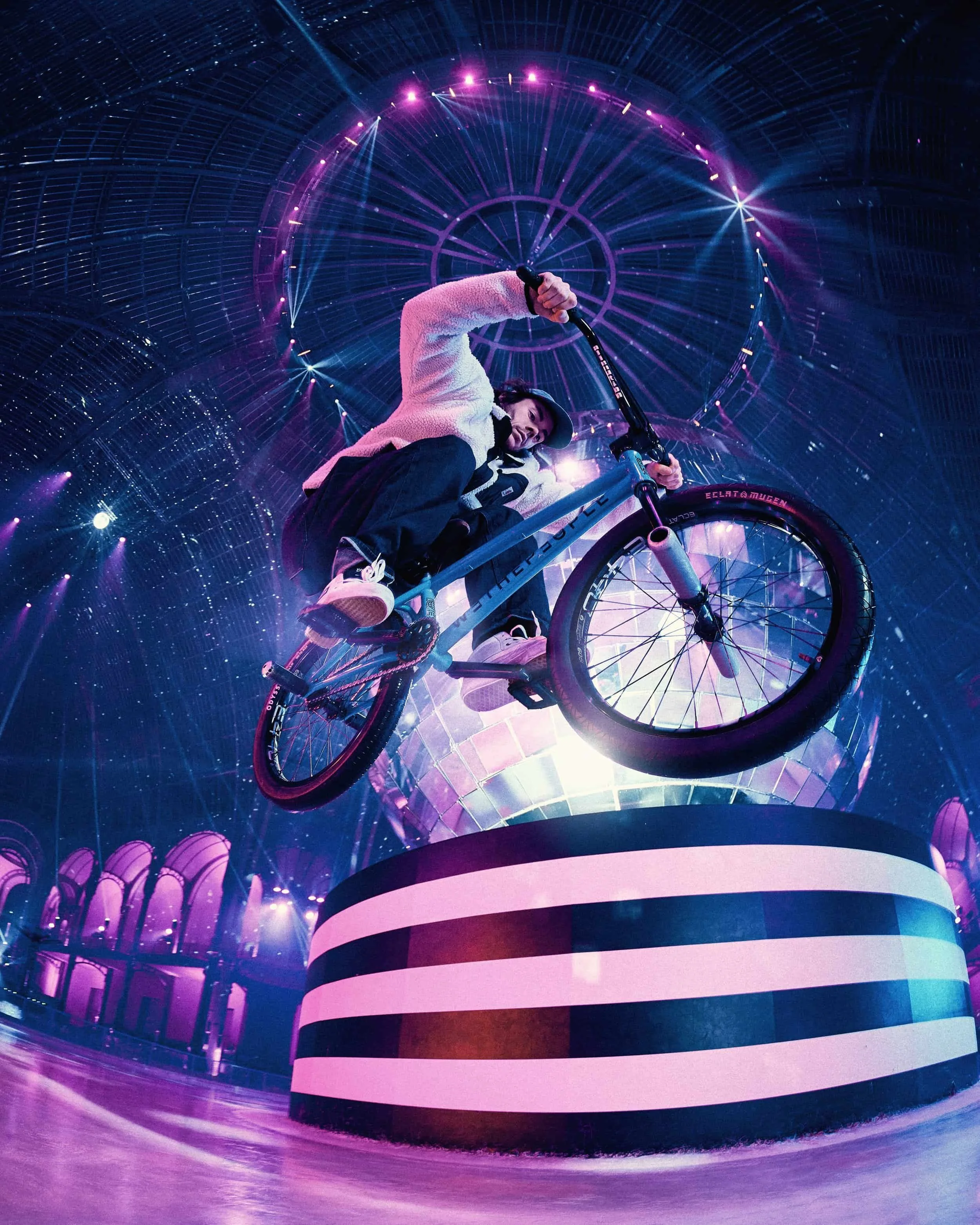 Un jeune homme en équilibre sur un vélo de BMX en plein saut dans un espace futuriste, avec un plafond éclairé de néons violets et bleus et un décor en forme de globe lumineux.