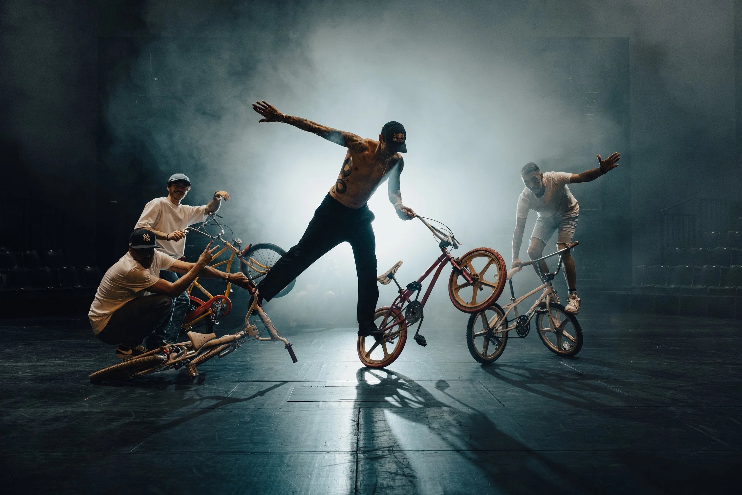 Groupe de quatre personnes avec des vélos, un de leurs membres est en train de faire une figure de BMX dans un bâtiment sombre avec des effets de fumée et de lumière en arrière-plan.