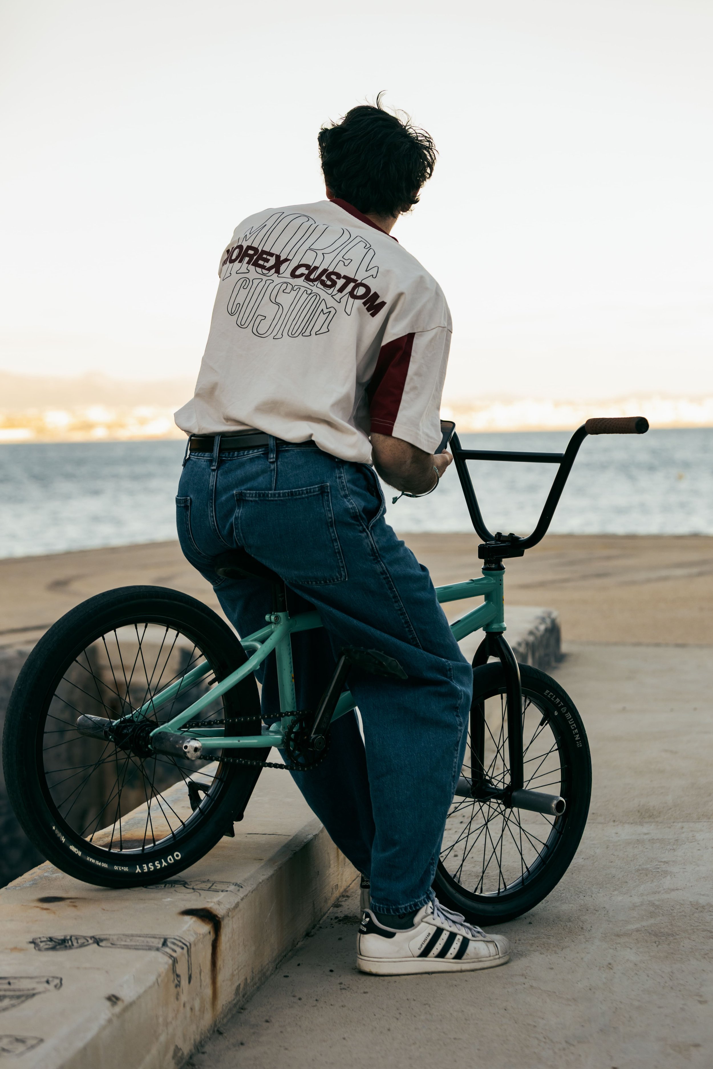 Rider de BMX flat assis sur son vélo, de dos, face à la mer au coucher de soleil à Marseille