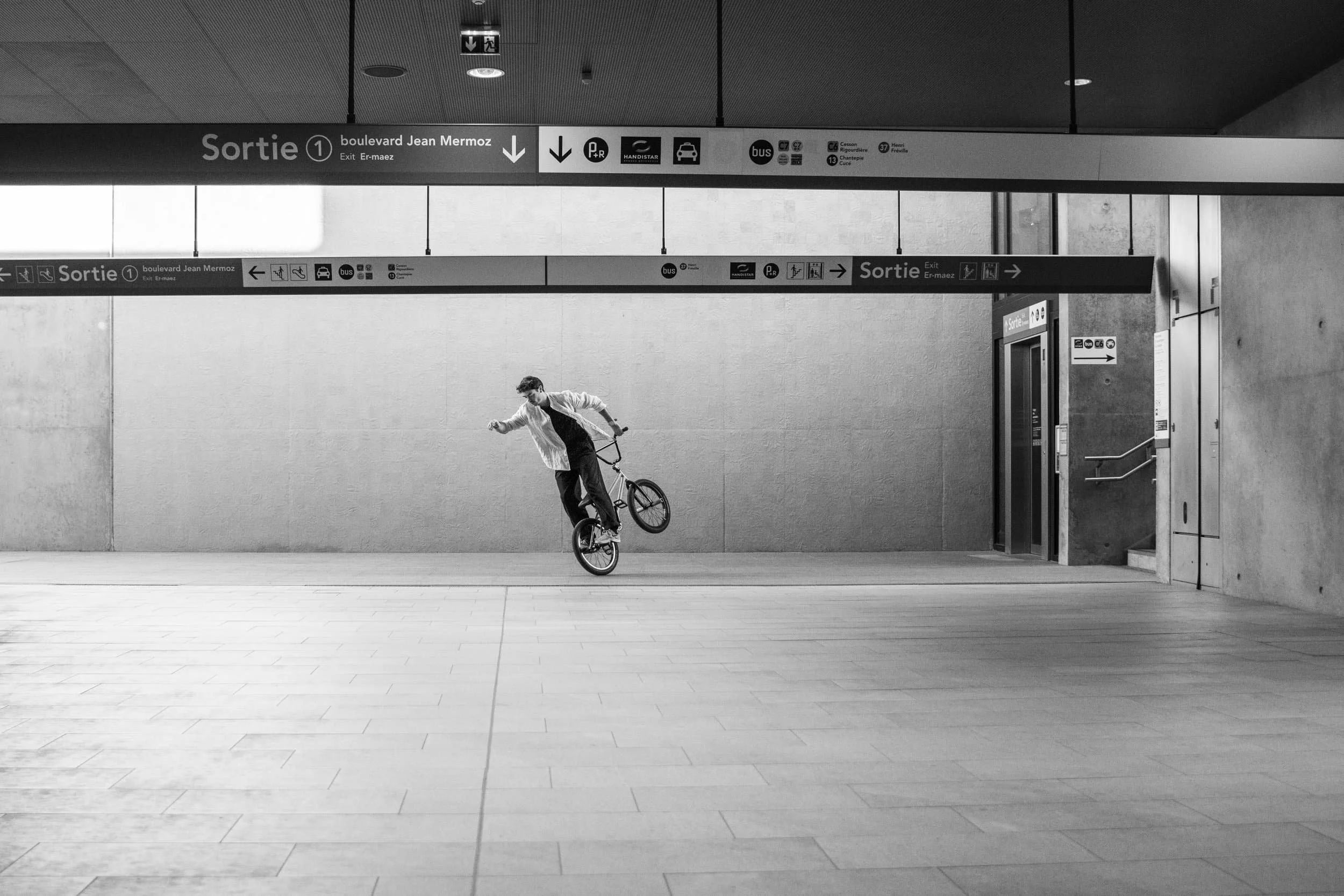 Un jeune homme faisant un seul wheelie sur un vélo dans une station de métro souterraine, en noir et blanc.