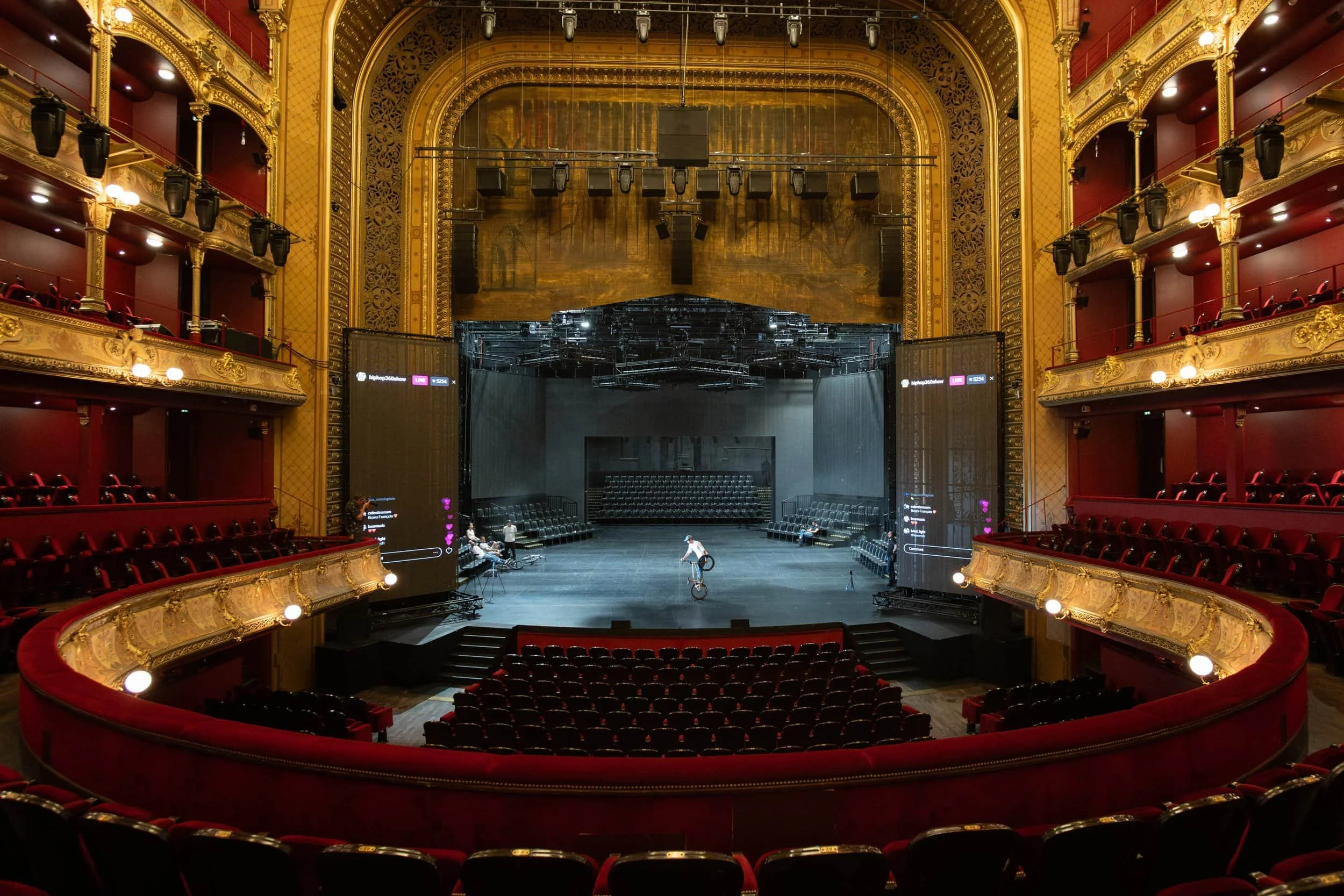 Une scène de théâtre ou de salle de spectacle vide avec un grand écran et une piste au centre, entourée de fauteuils rouges et de balcons ornés de dorures, dans une salle élégante et luxueuse.
