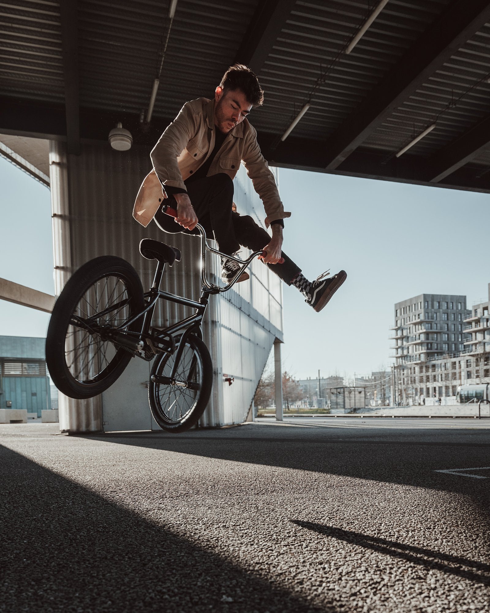 Un jeune homme effectue une figure en BMX sous un pont, avec des bâtiments modernes en arrière-plan.