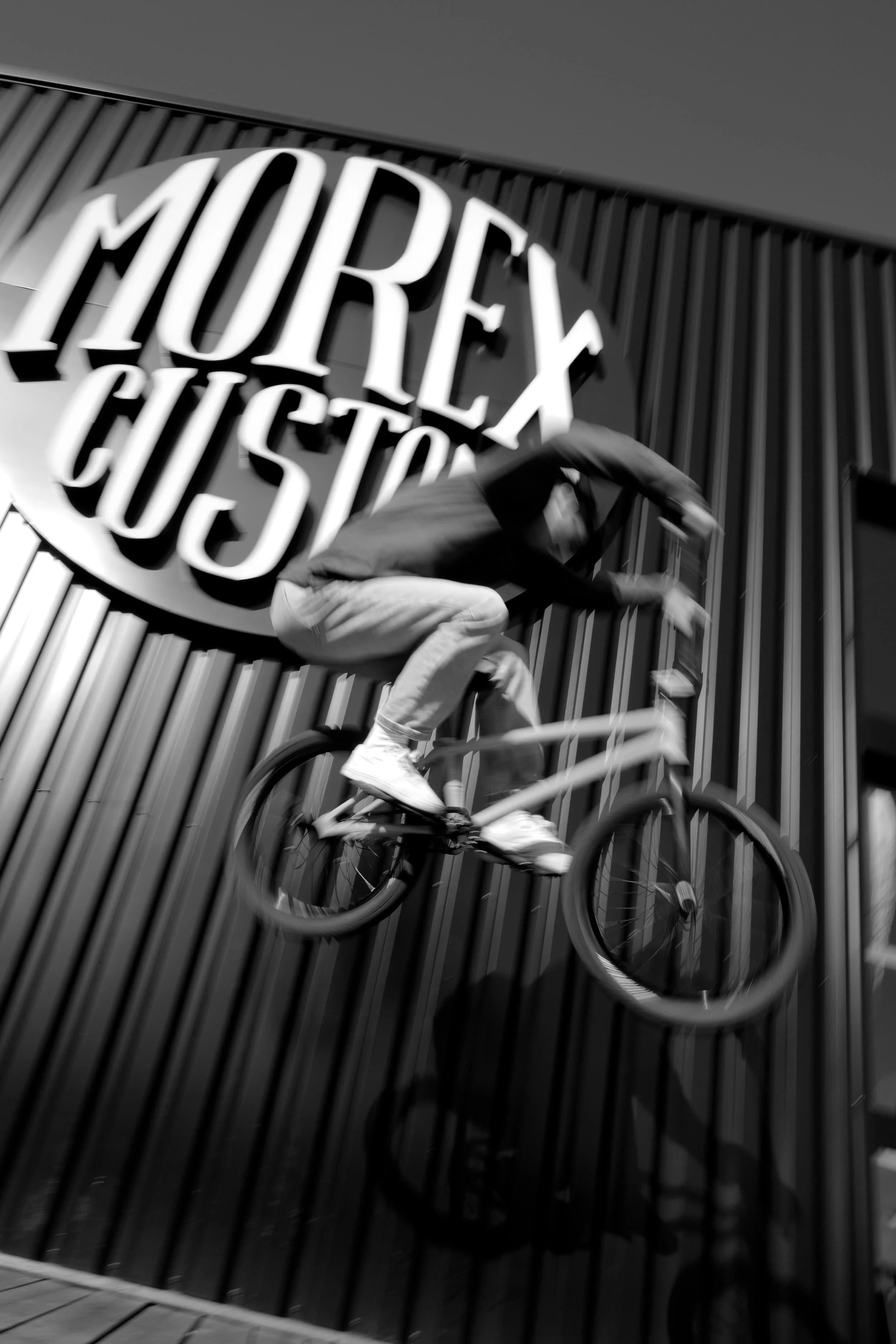 Un homme faisant du BMX en accélération devant un mur avec un grand logo 'Morex Custom'.