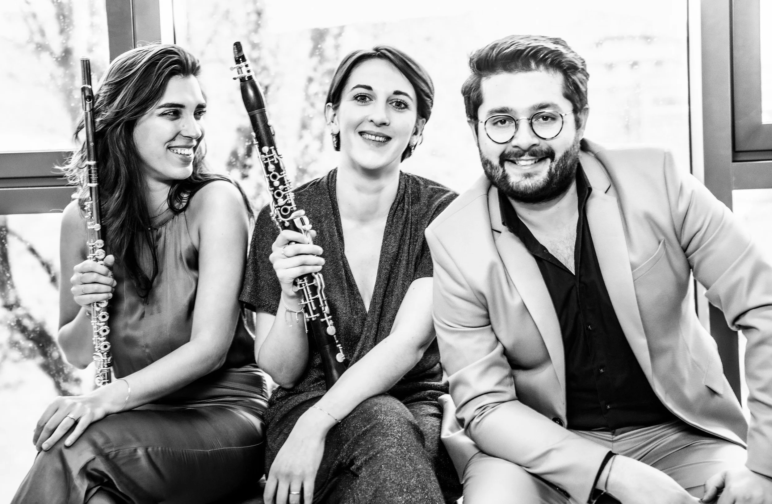 Three musicians holding clarinets sit together near a window, smiling at the camera.