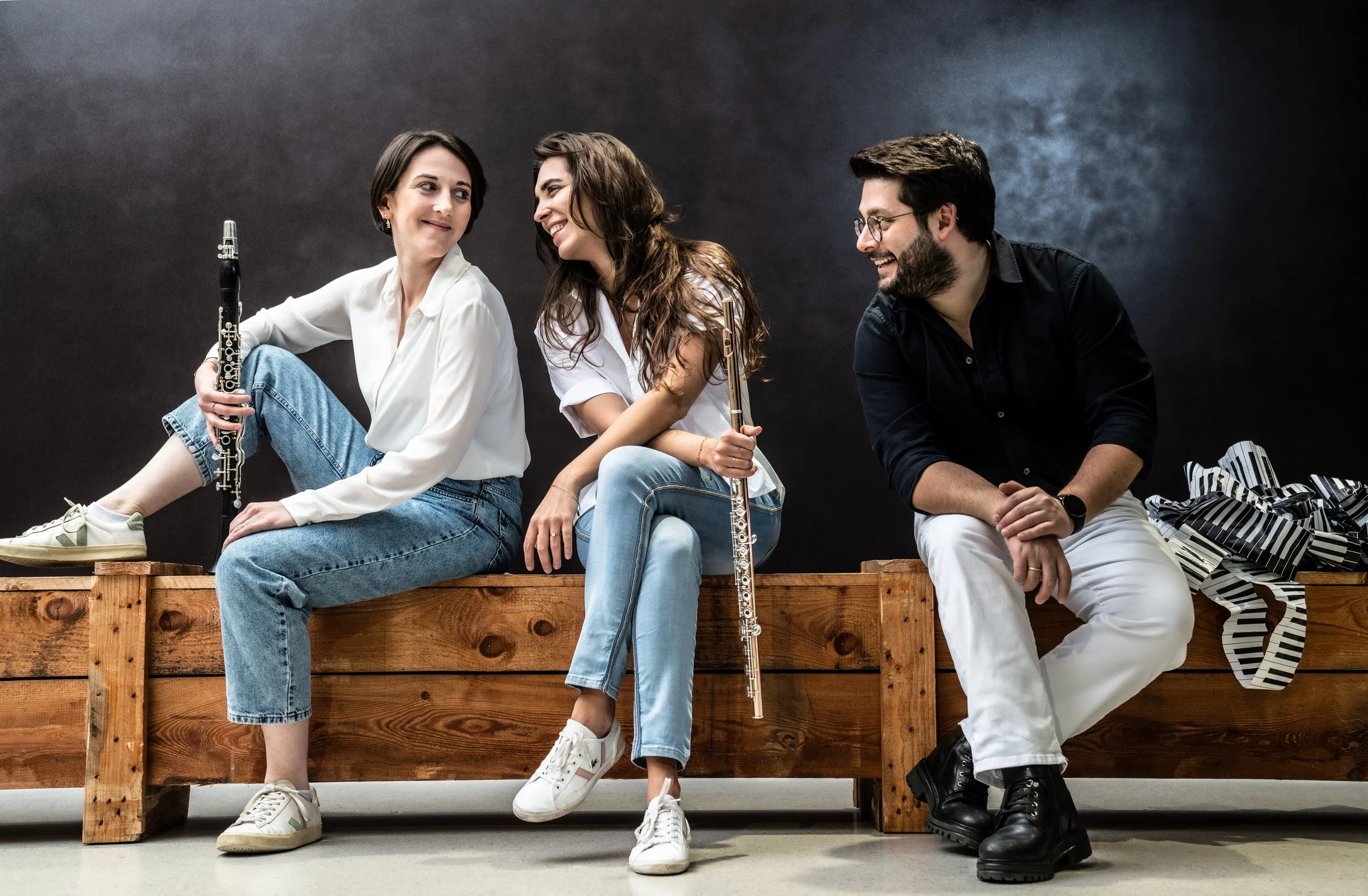 Three people sitting on a wooden bench, two women holding flutes, a man sitting beside them, all smiling and engaging in conversation against a dark background, with wrapped gifts nearby.