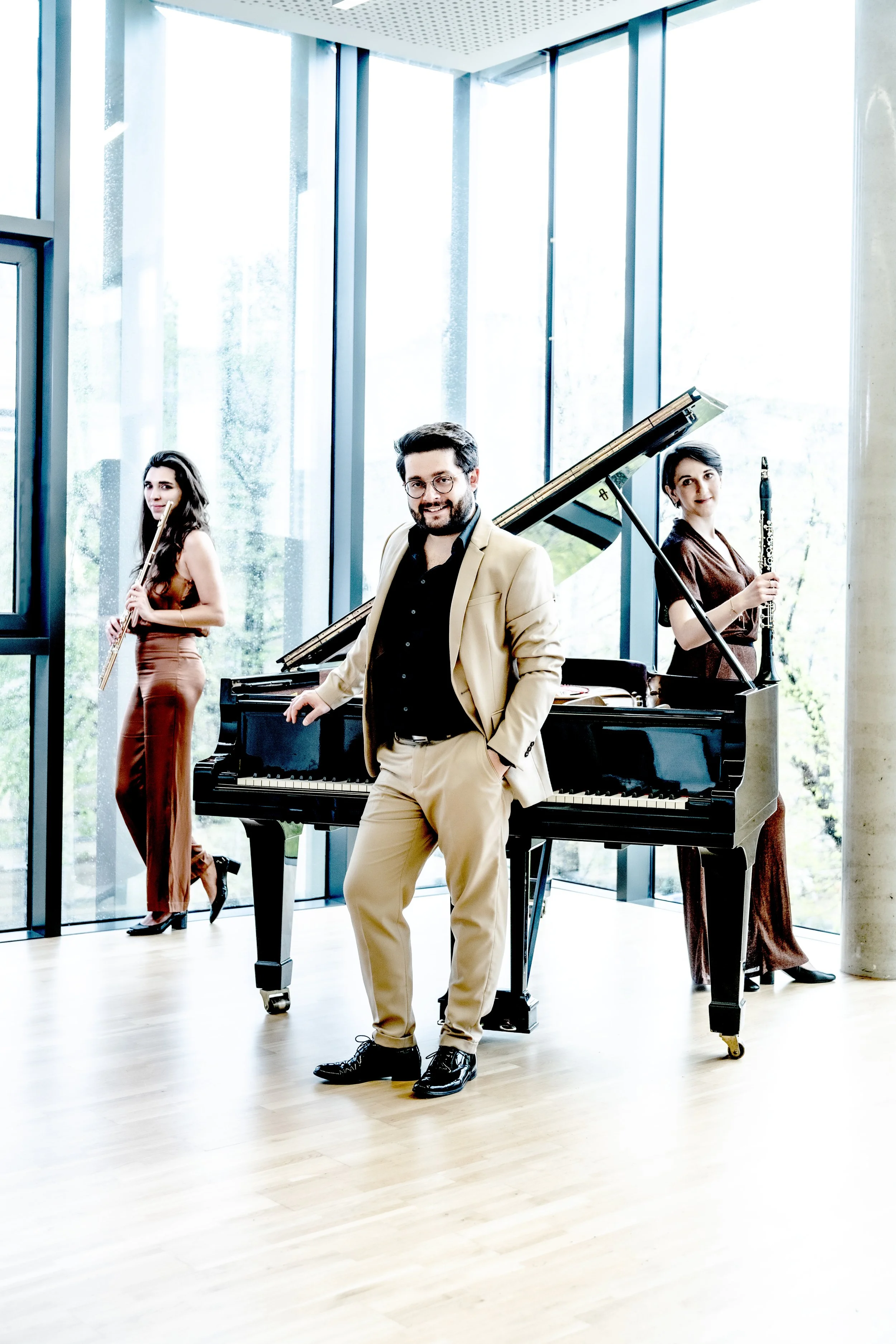 Three musicians in a modern, sunlit room: a man in a light beige suit standing in front of a black grand piano, a woman in a brown dress holding a clarinet standing behind him, and another woman in a reddish-brown dress holding a flute standing to th