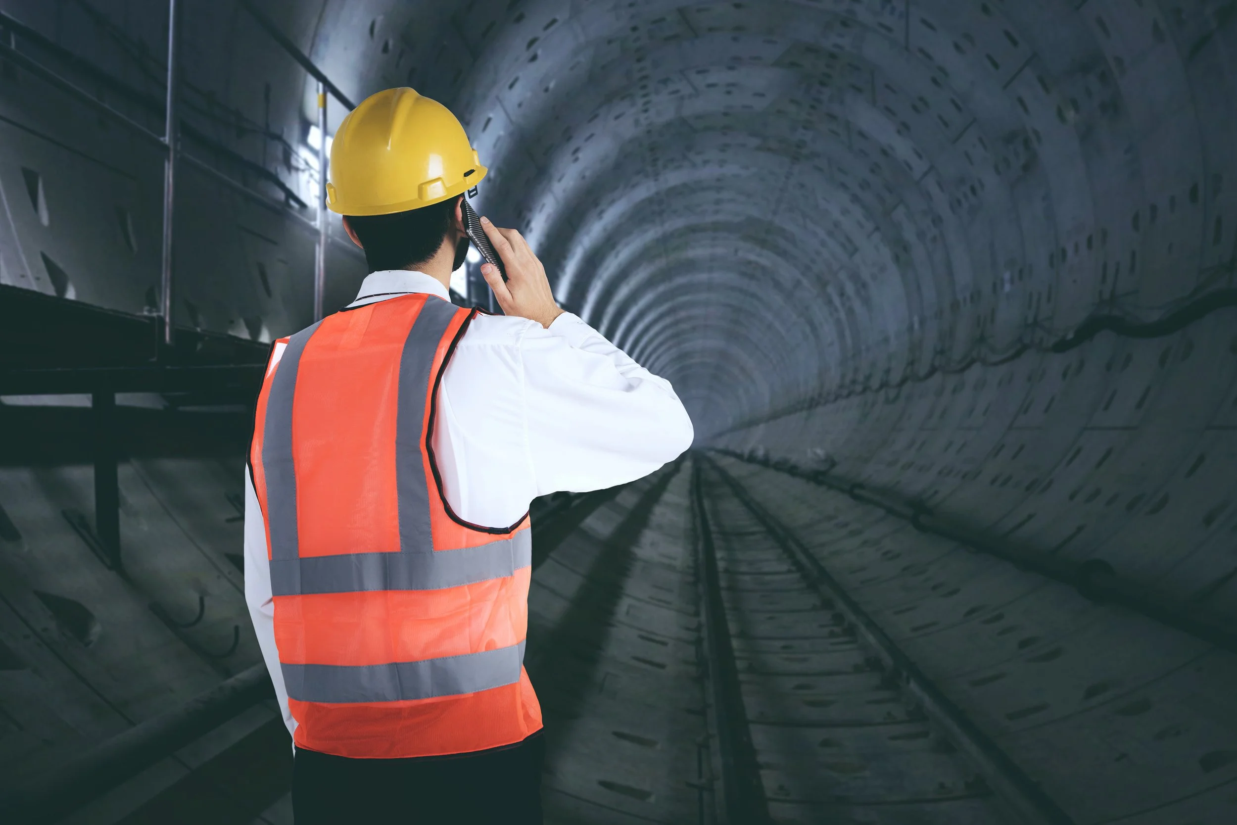 Bauarbeiter mit gelbem Schutzhelm und orangefarbener Warnweste telefoniert in einem Tunnel.
