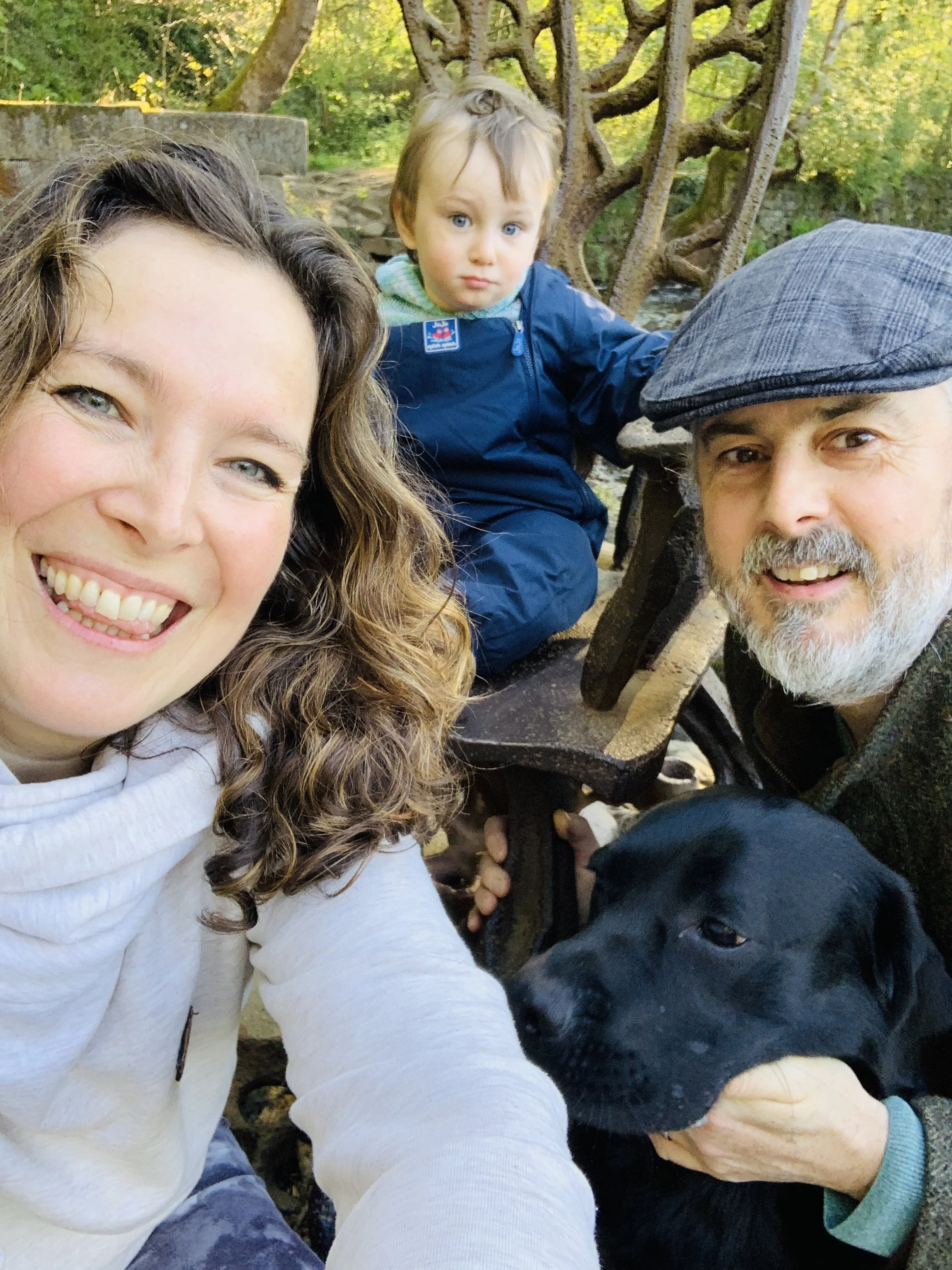 A family outdoor selfie with a smiling woman, a young boy with serious expression, a man with a beard and cap, and a black dog sitting on a park bench with trees in the background.