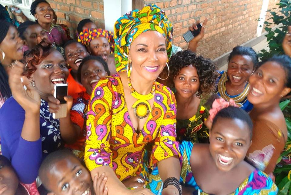 Group of women and girls smiling and posing for a photo outdoors, some wearing colorful traditional African clothing and headwraps.