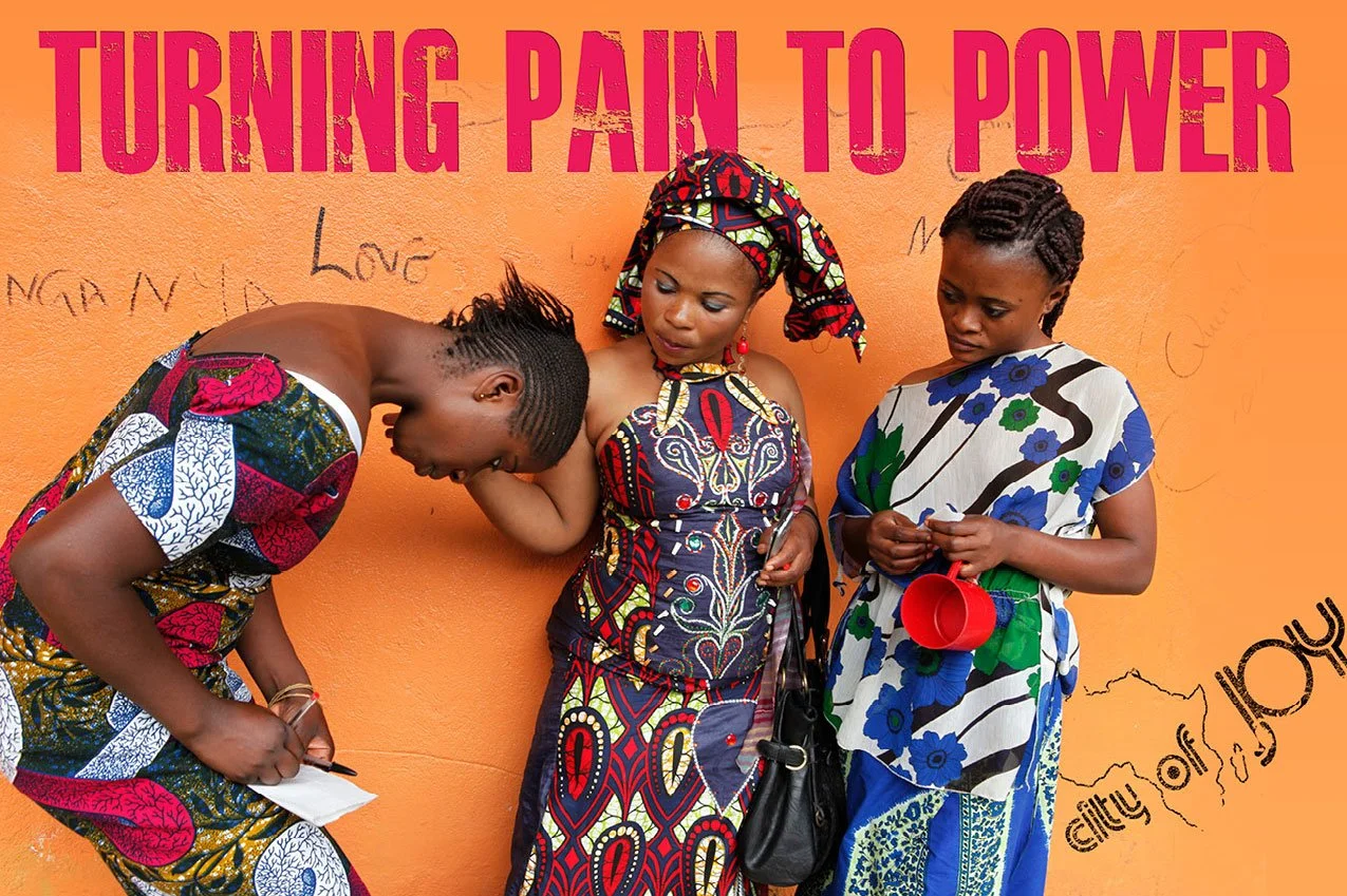 Three women in colorful African dresses standing against an orange wall with handwritten messages. They appear to be in conversation, focused and engaged. There is large pink text above them that reads 'Turning Pain to Power'.