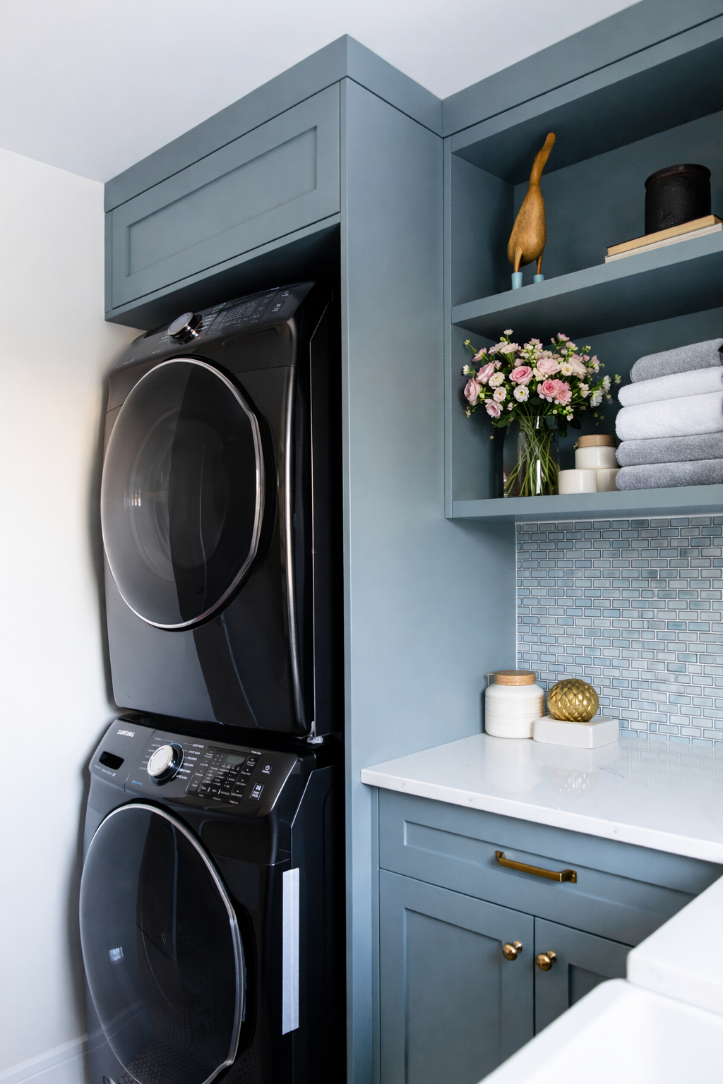 A laundry room with a stacked black washing machine and dryer, blue cabinets, a white countertop, and decorative items including a vase with pink flowers, folded towels, candles, and ornamental objects.
