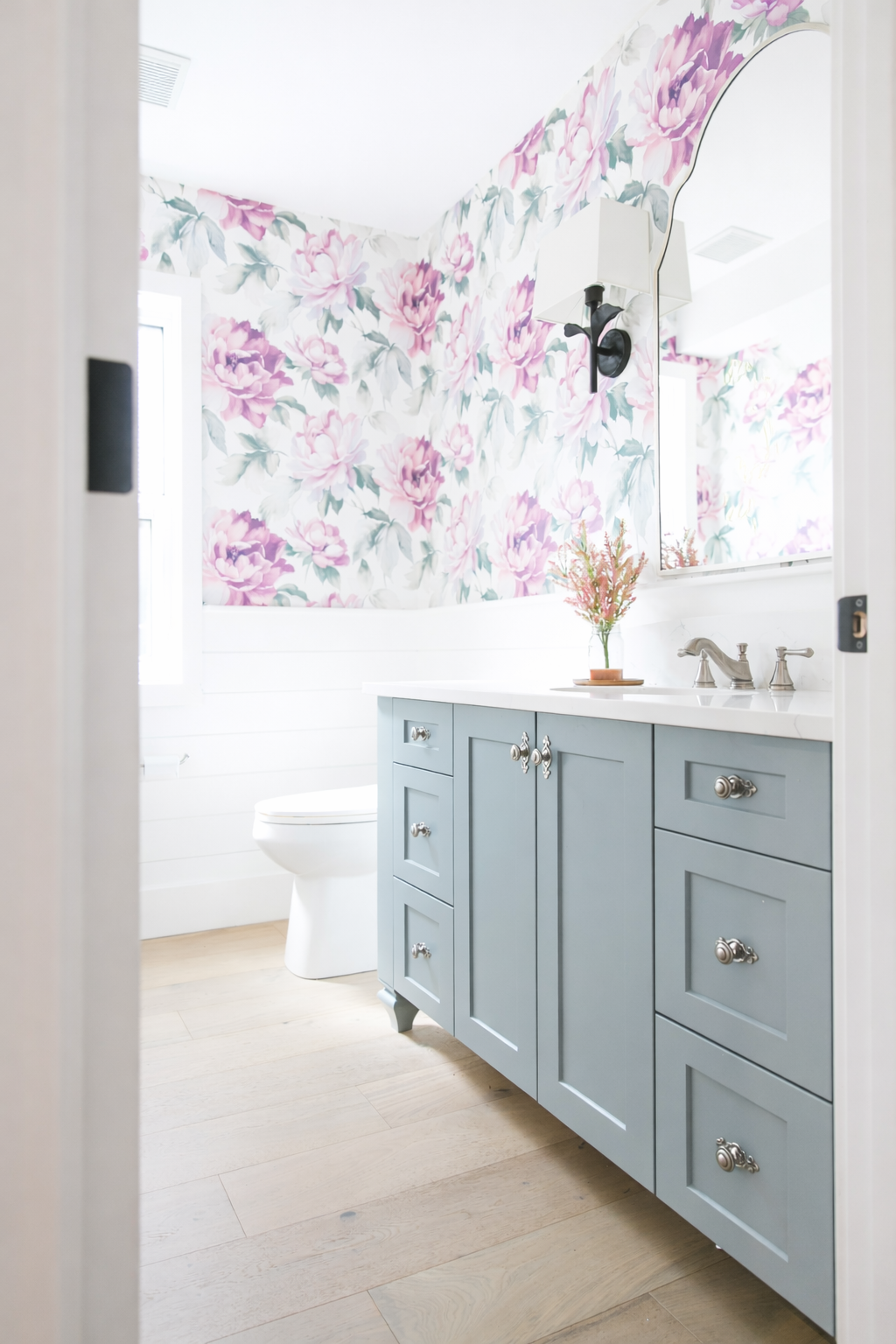 A bathroom with floral wallpaper, a blue vanity with silver knobs, a white countertop, a small potted plant in pink flowers, and a mirror above the sink. The bathroom has a white toilet, hardwood flooring, and natural light from a window.