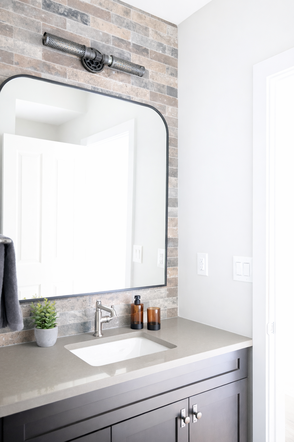 Modern bathroom vanity with a gray cabinet, beige countertop, rectangular sink, silver faucet, a small potted plant, and brown soap dispenser and matching cup. A large mirror is mounted on a brick wall, with a ventilation pipe above.
