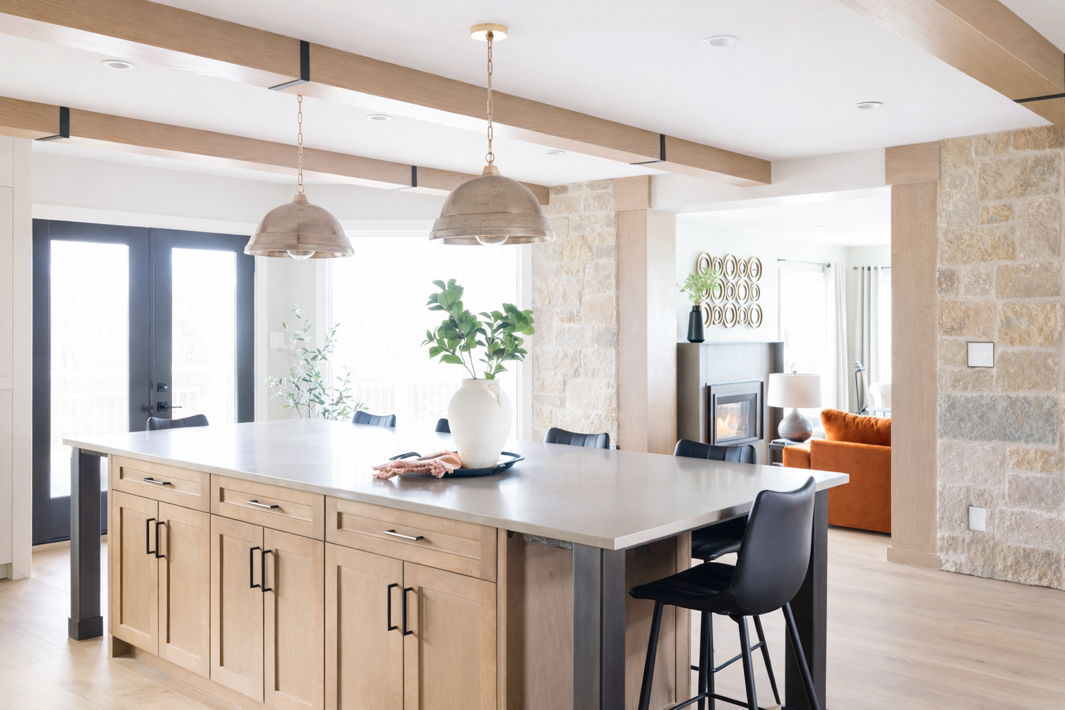 Modern open-plan kitchen with a large island, black chairs, hanging pendant lights, and a view of the living area with an orange sofa and stone accents.