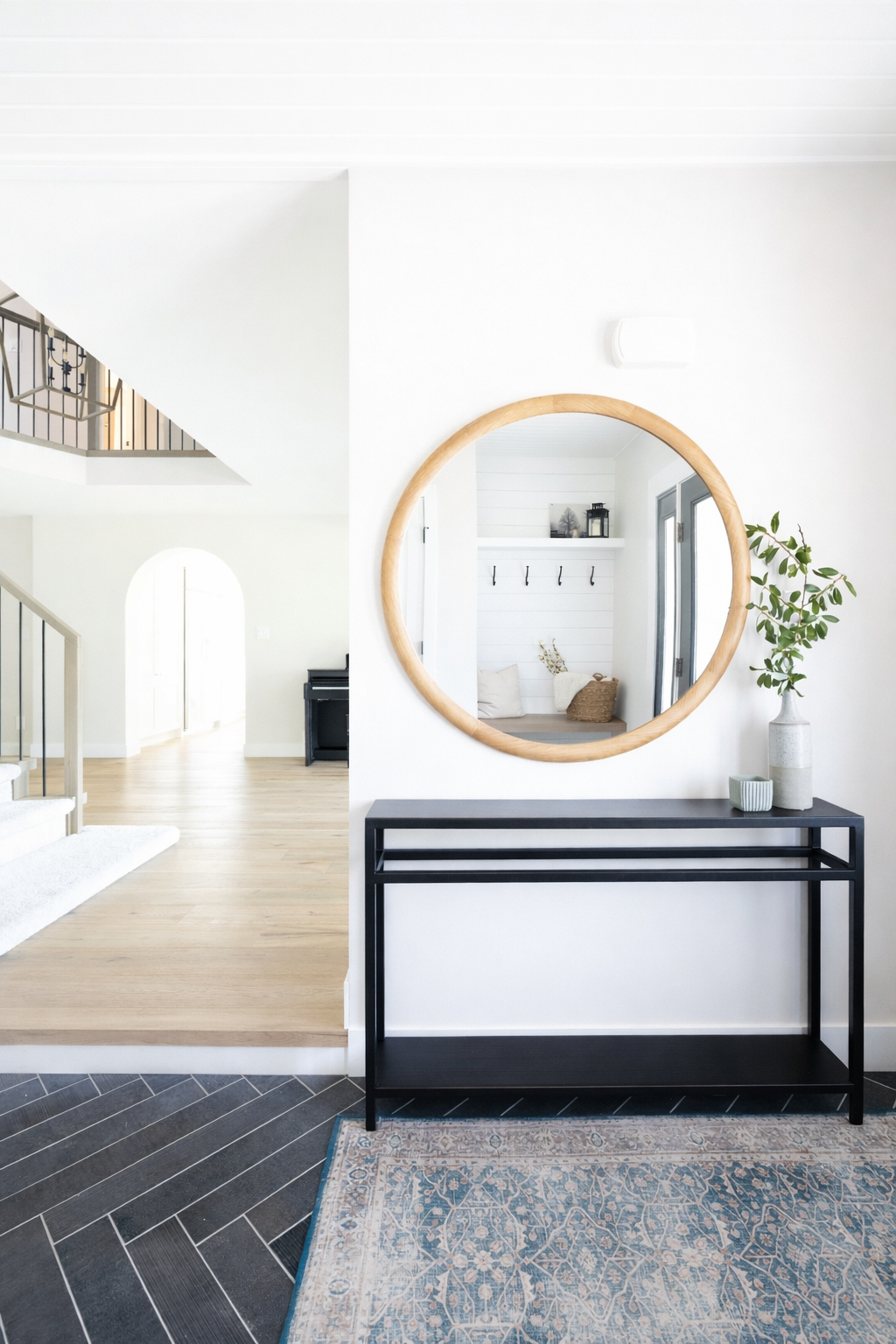 Modern interior entryway with a black console table, a round wooden-framed mirror, a large white vase with green branches, and a small patterned container. The space is bright with white walls and light wood flooring.