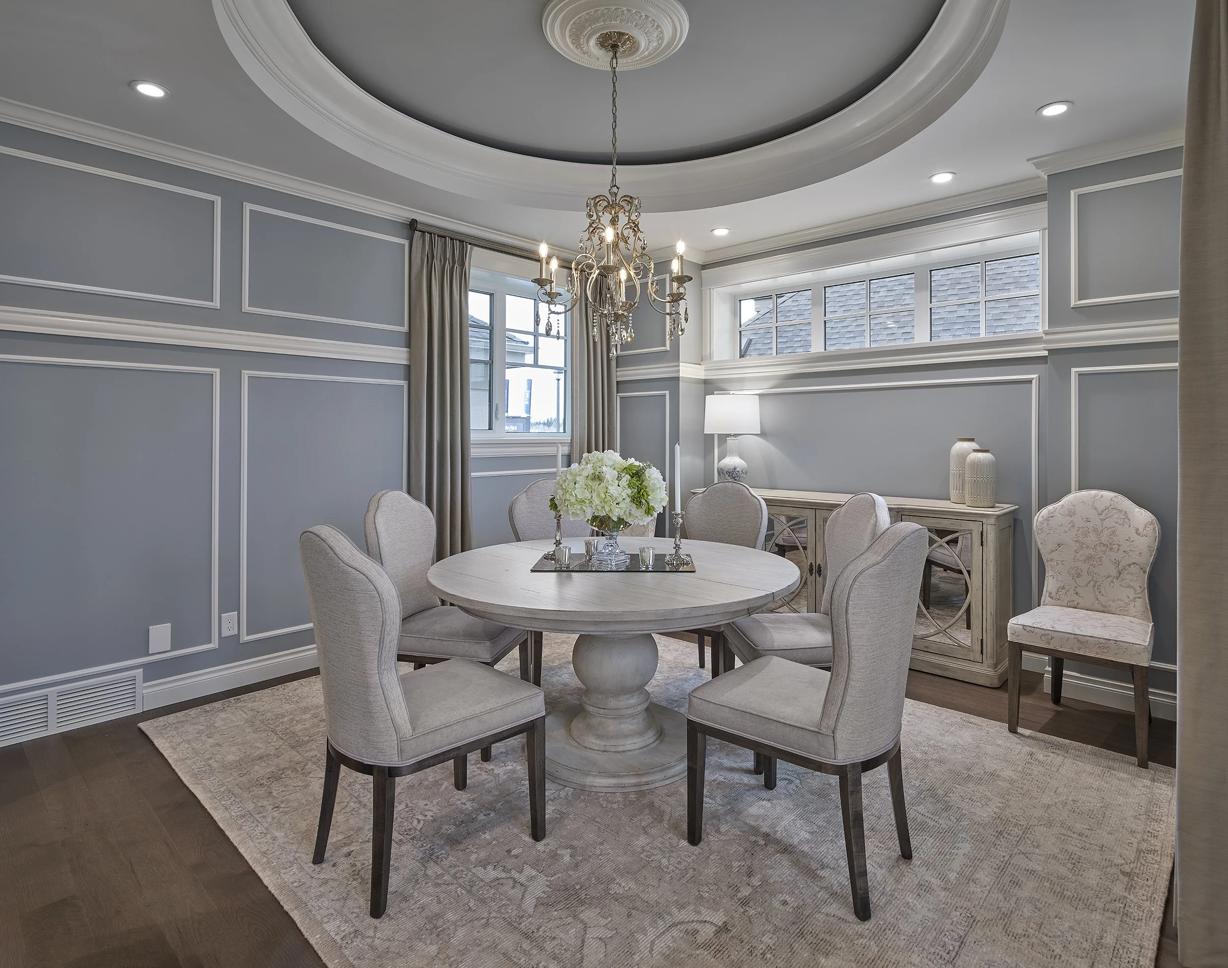 Elegant dining room with a round table surrounded by six upholstered chairs, a chandelier hanging above, and a sideboard with decorative vases. Light blue walls with white trim, windows with beige curtains, and hardwood flooring with a beige rug.