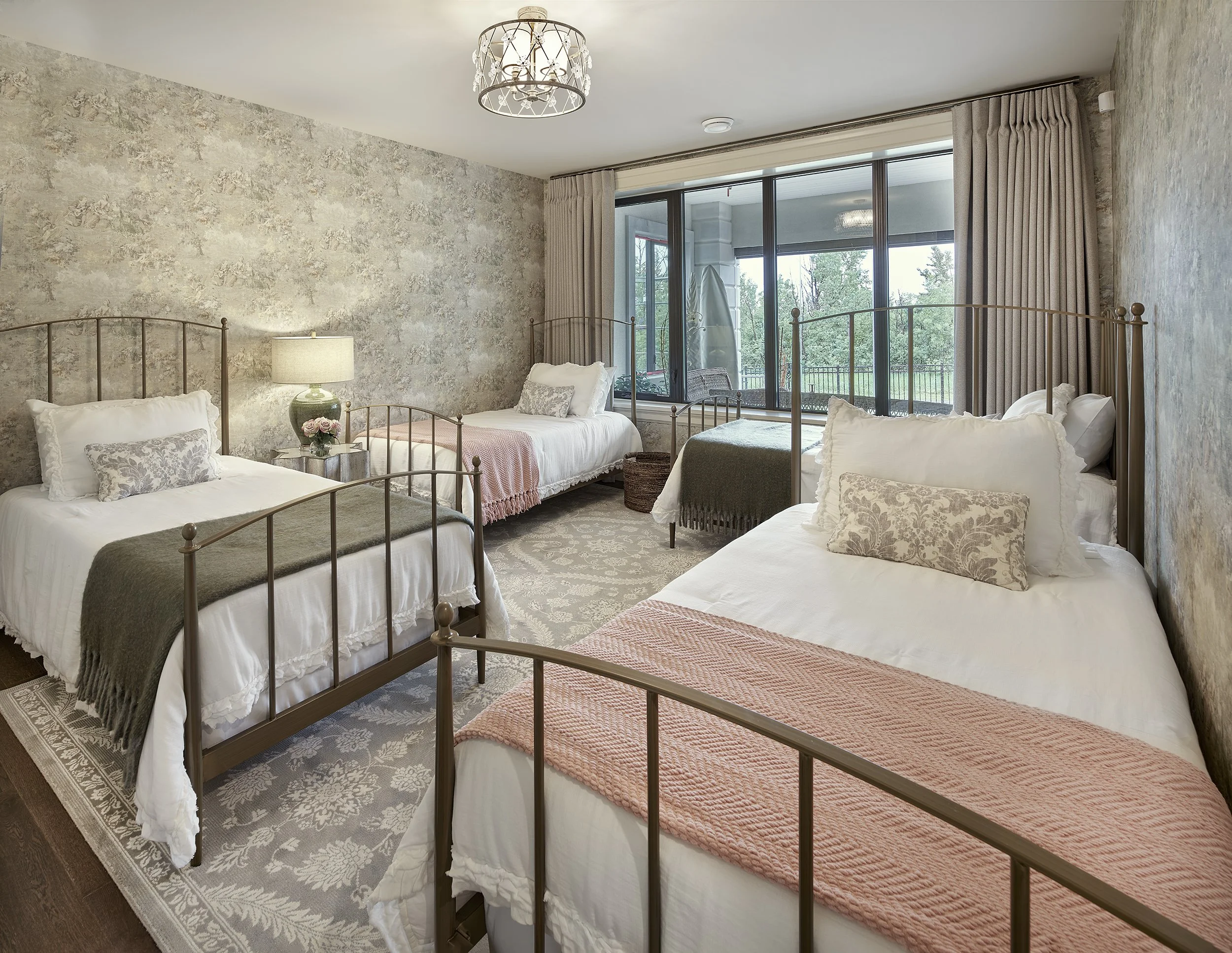 A bedroom with four metal frame beds, each with floral pillows, blankets, and white bedding. Large window with curtains shows trees outside. There is a bedside table with a lamp and small floral arrangement between two beds, and a sitting area beyond the window.