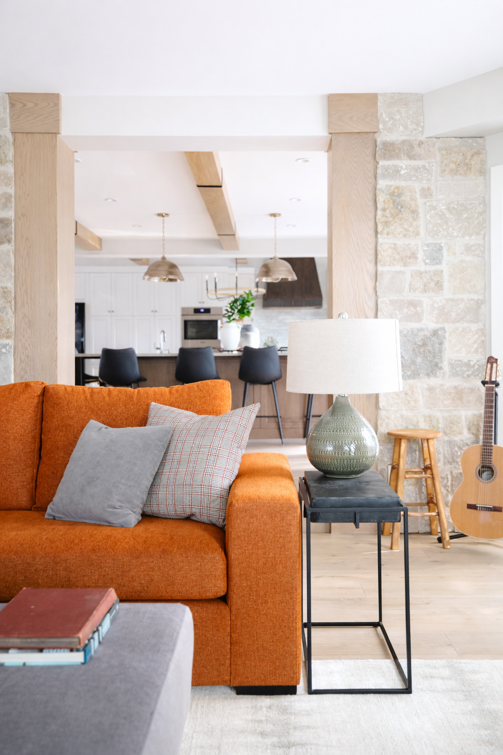 Living room with orange sofa, gray pillows, a side table with a green ceramic lamp, a guitar, and a stone wall. Kitchen in the background with black bar stools and a dining area.