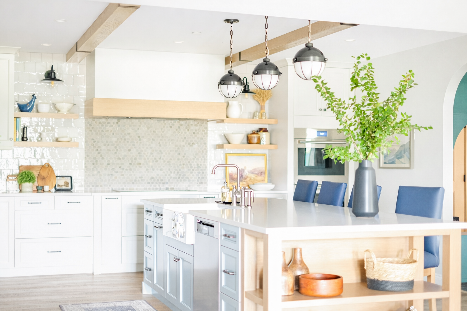 Modern kitchen with white cabinets, island with blue chairs, large vase with green branches, pendant lights, and open shelving with decorative items.