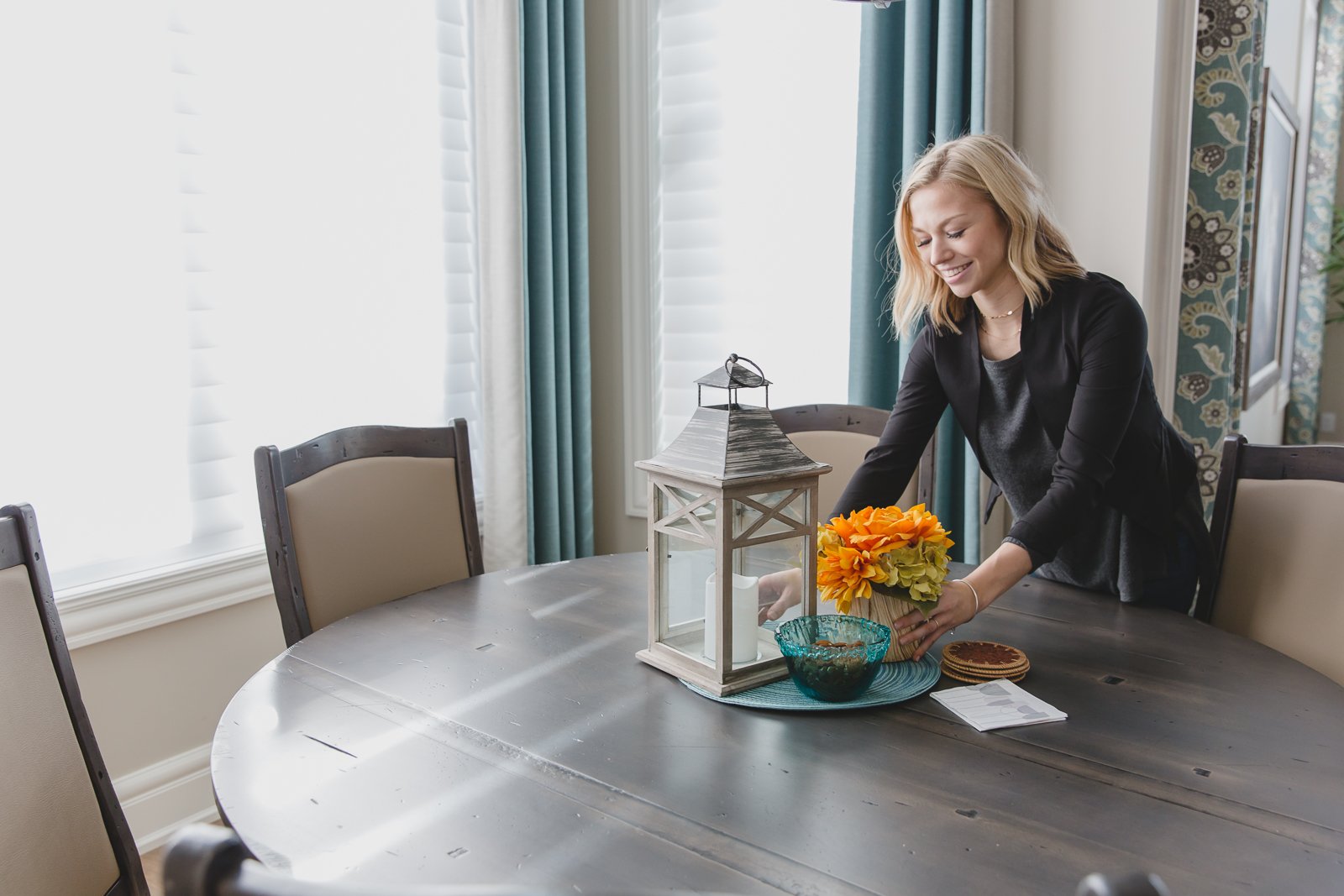 A woman is placing a flower arrangement on a dining table with a black finish, surrounded by beige and dark wood chairs, in a bright room with light blue and white curtains and decorative wallpaper.