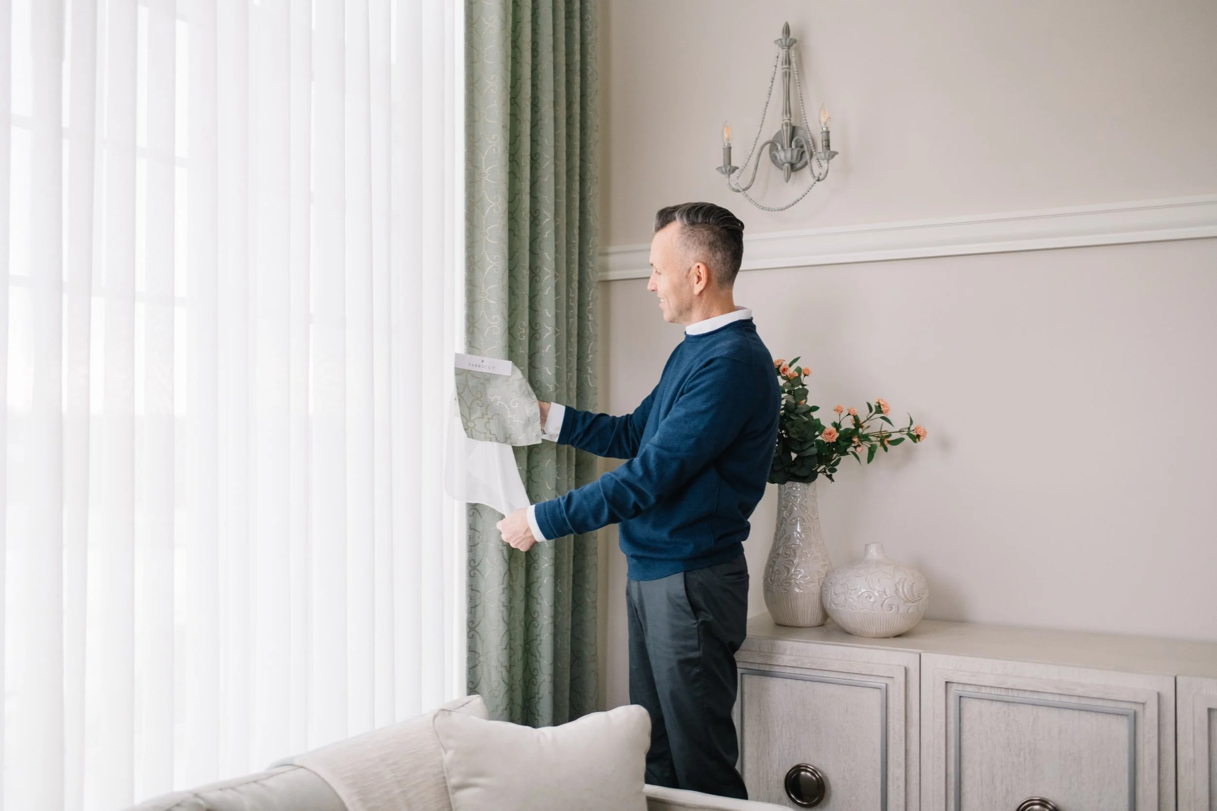 A man with gray hair and a side shave, wearing a blue sweater and dress shirt, opening a curtain in a living room with cream walls, green curtains, and decorative vases.
