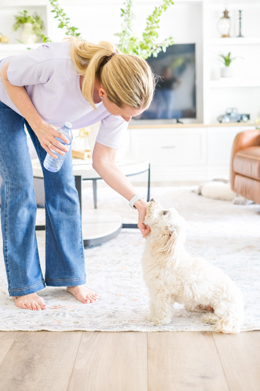 Woman playing with a small white puppy indoors, sunlight, modern living room, cozy sofa, decorative items.