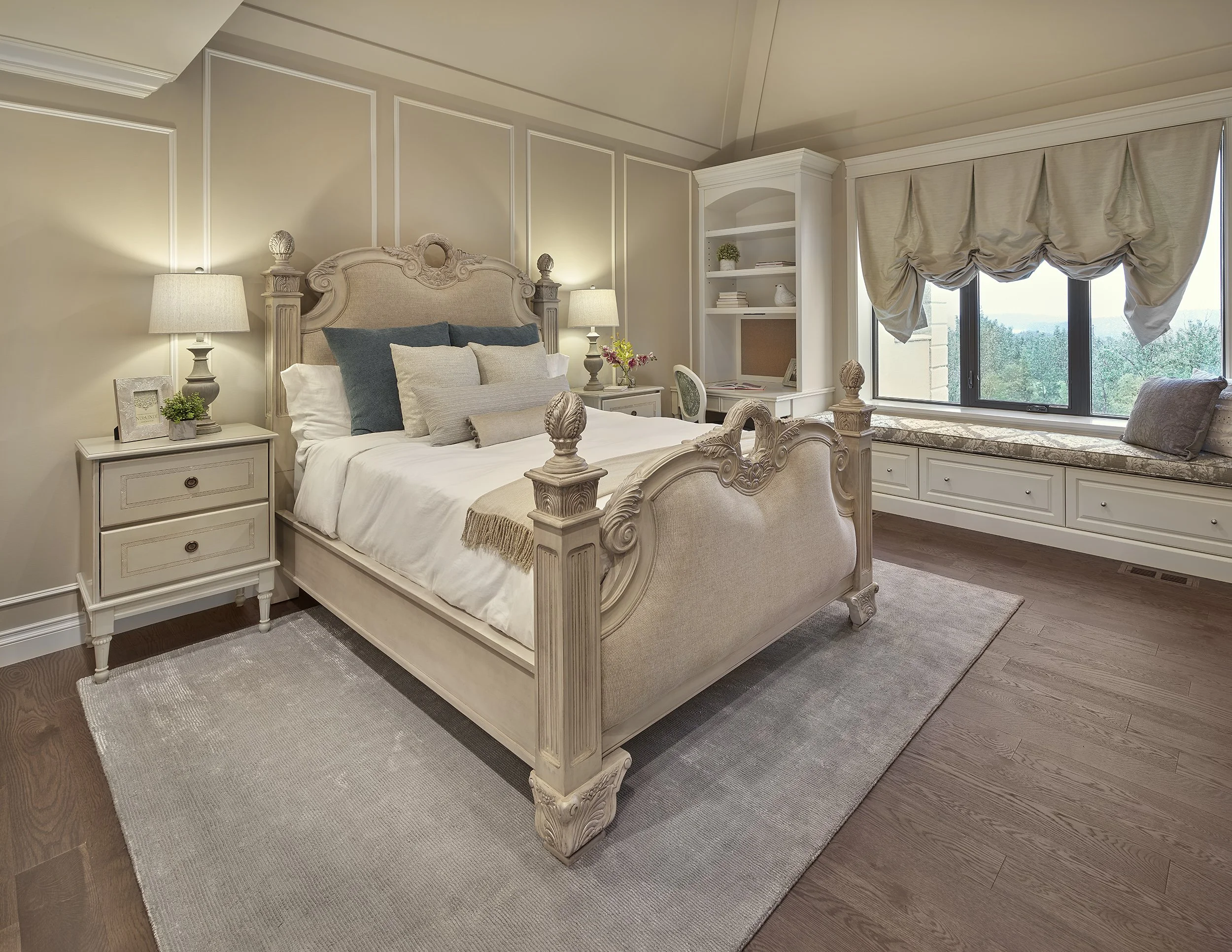 A cozy bedroom with a large ornate cream-colored bed, matching nightstands with lamps, a built-in window seat with decorative pillows, and a white bookshelf. Bright natural light fills the room through a large window with cream drapes, and a beige ar